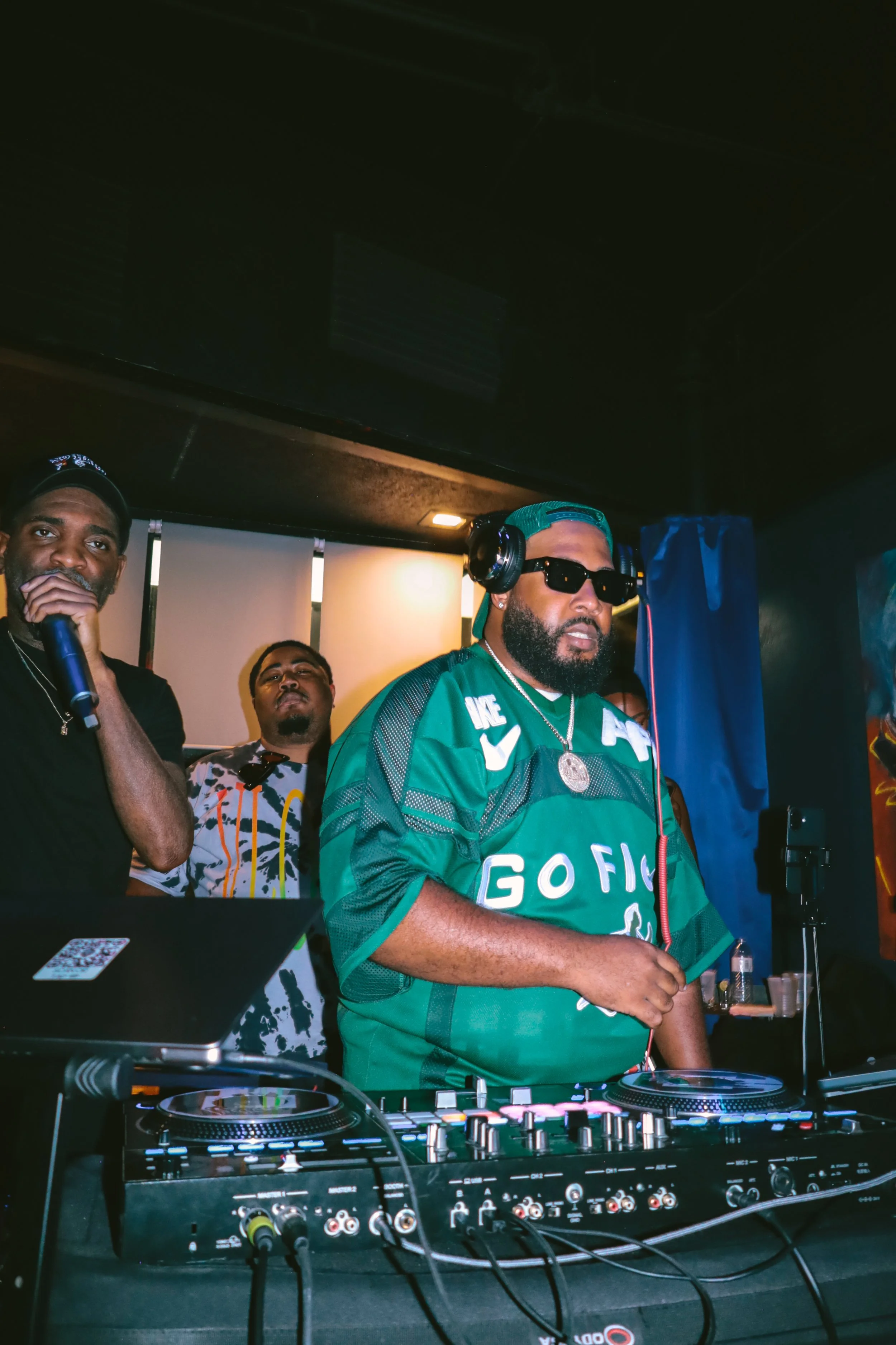 A DJ wearing a green shirt, sunglasses, and a turquoise cap, spinning music with turntables and a mixer at a club event, surrounded by people.