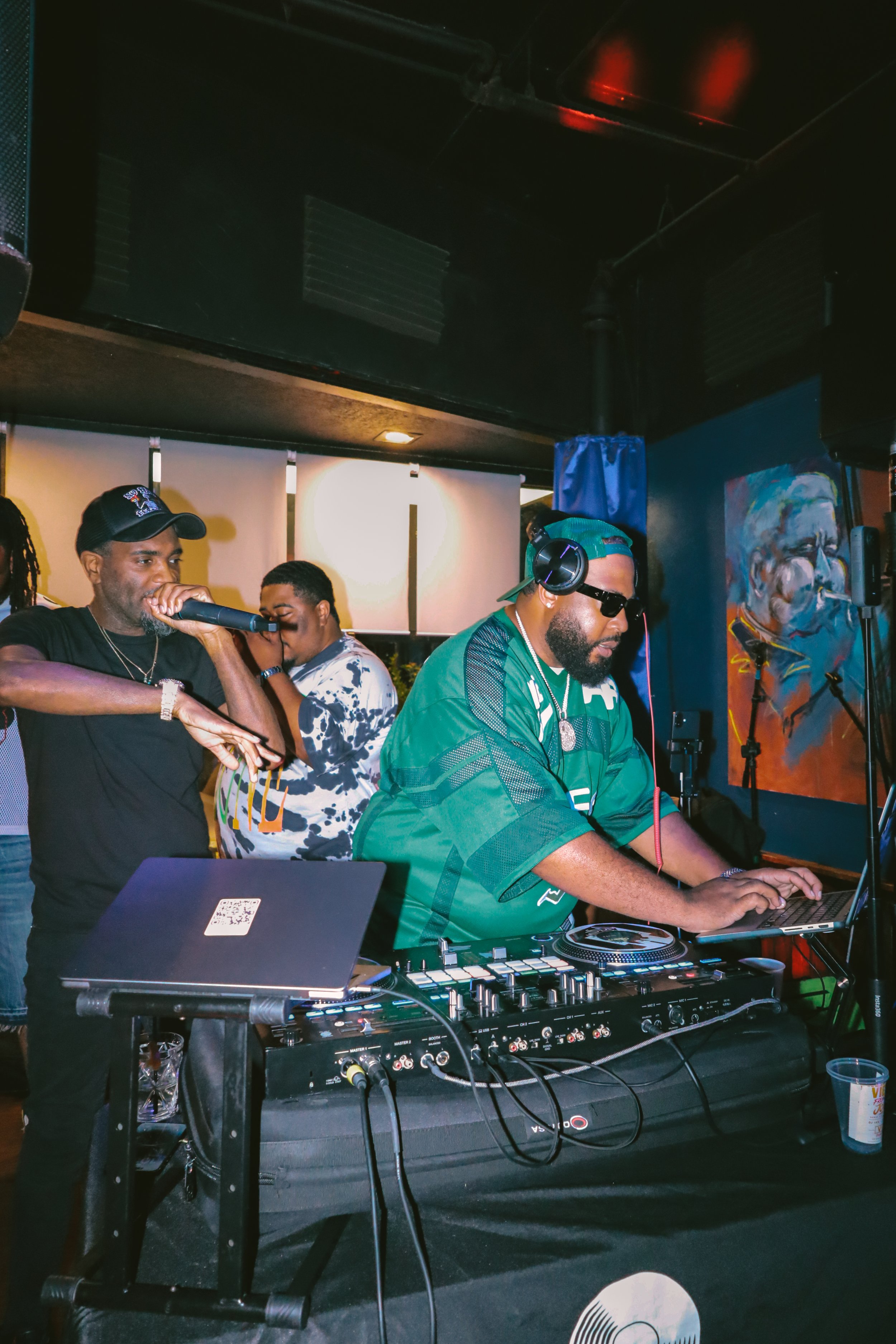 A DJ wearing a green jersey and headphones is operating DJ equipment with a laptop nearby. Two men are standing behind him, one holding a microphone and the other looking on. The setting appears to be an indoor event or party with colorful artwork on