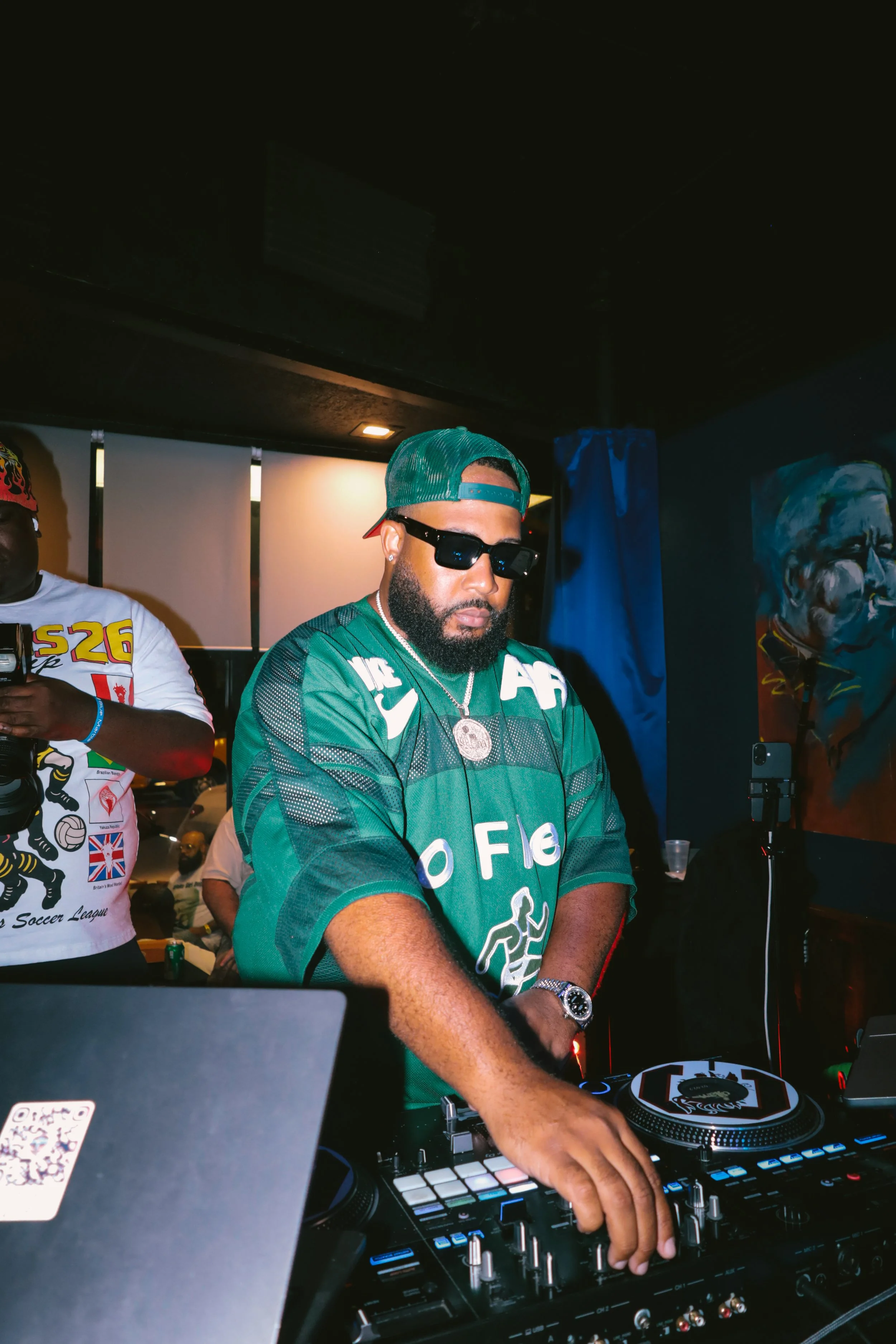A DJ with a beard wearing sunglasses, a green sports jersey, a green cap backward, and jewelry, is mixing music on a DJ controller at a social event. To his left, another man in a white t-shirt with colorful graphics is holding a drink and a phone. T