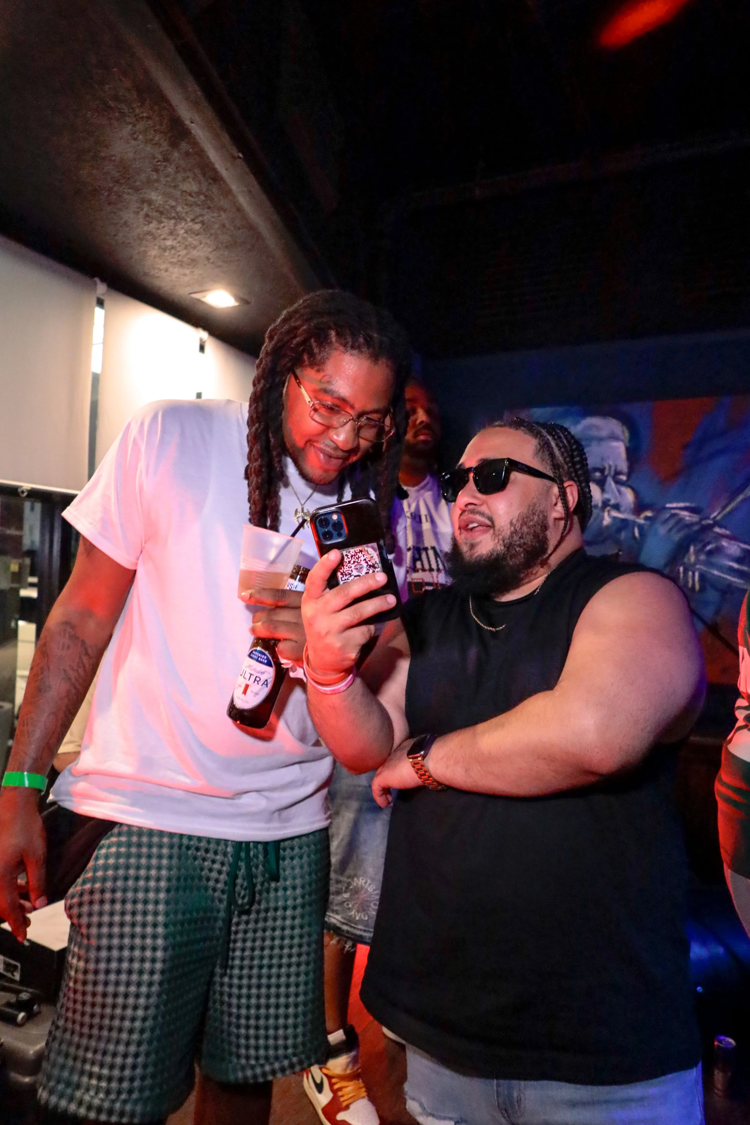Two men are taking a selfie at a party or social gathering. One man is wearing glasses, a white T-shirt, and patterned shorts, holding a drink and a phone. The other man, in black sunglasses, a sleeveless black shirt, and gray pants, is sitting and s