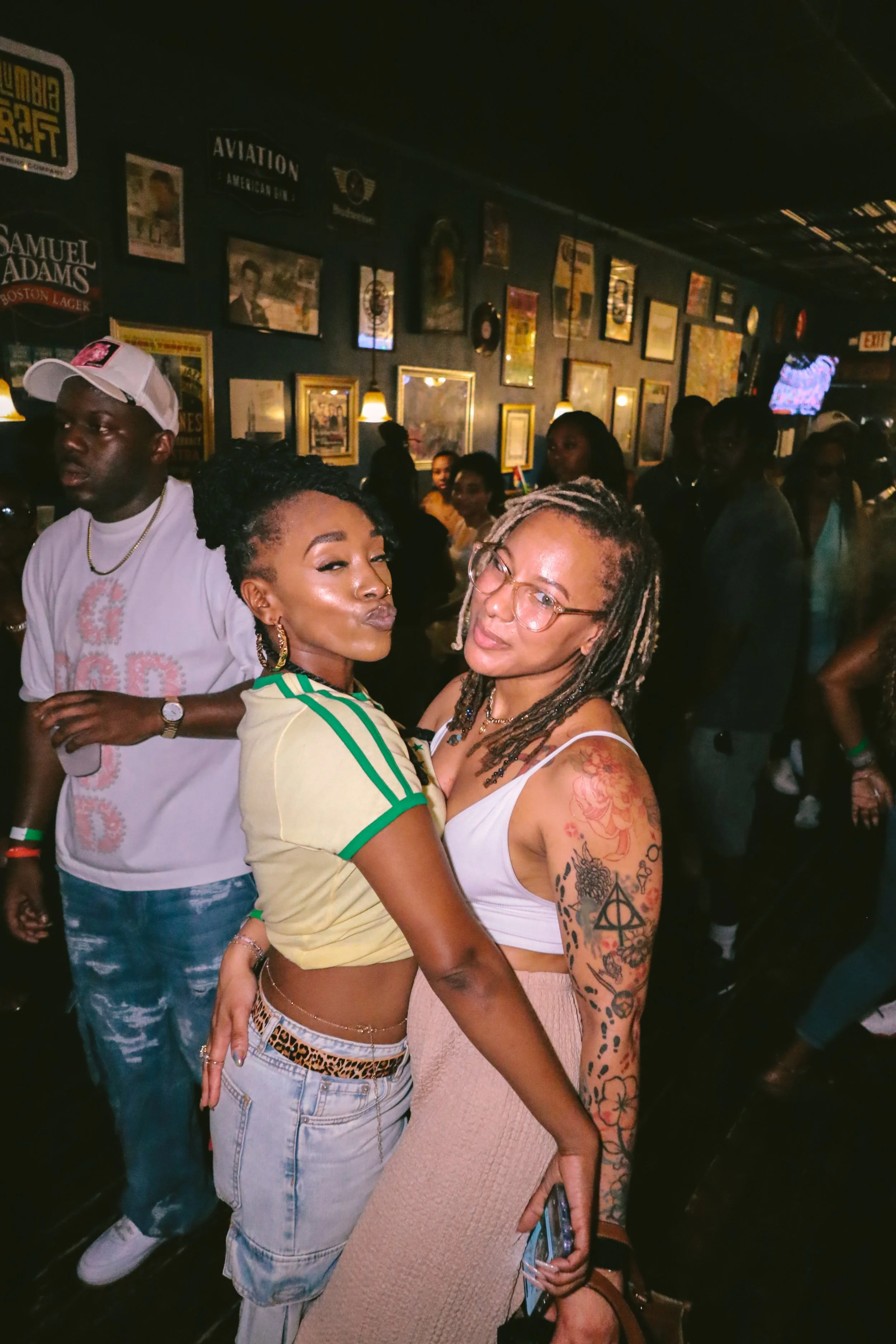 Two women kissing and hugging in a crowded bar or club, with people socializing in the background, decorated with framed pictures and signs on the wall.