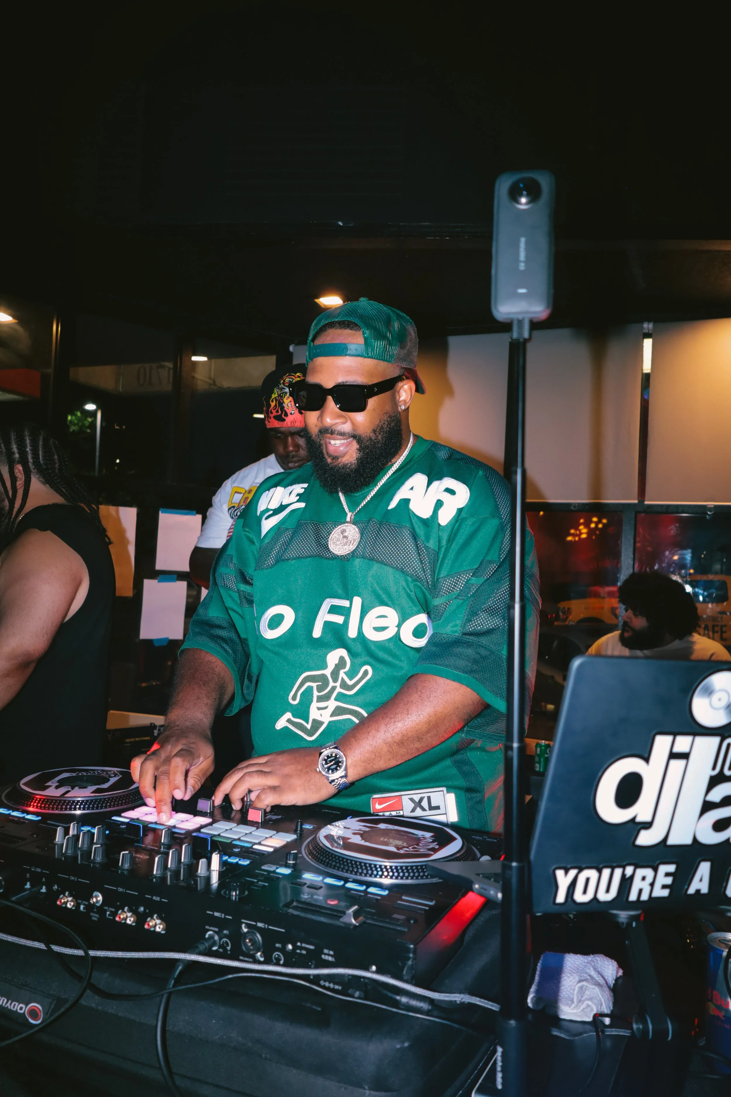 A DJ wearing sunglasses and a green Nike football jersey is DJing with a mixer and turntables in a dimly lit indoor space, with a person in the background wearing a white shirt with a fiery design on the head.