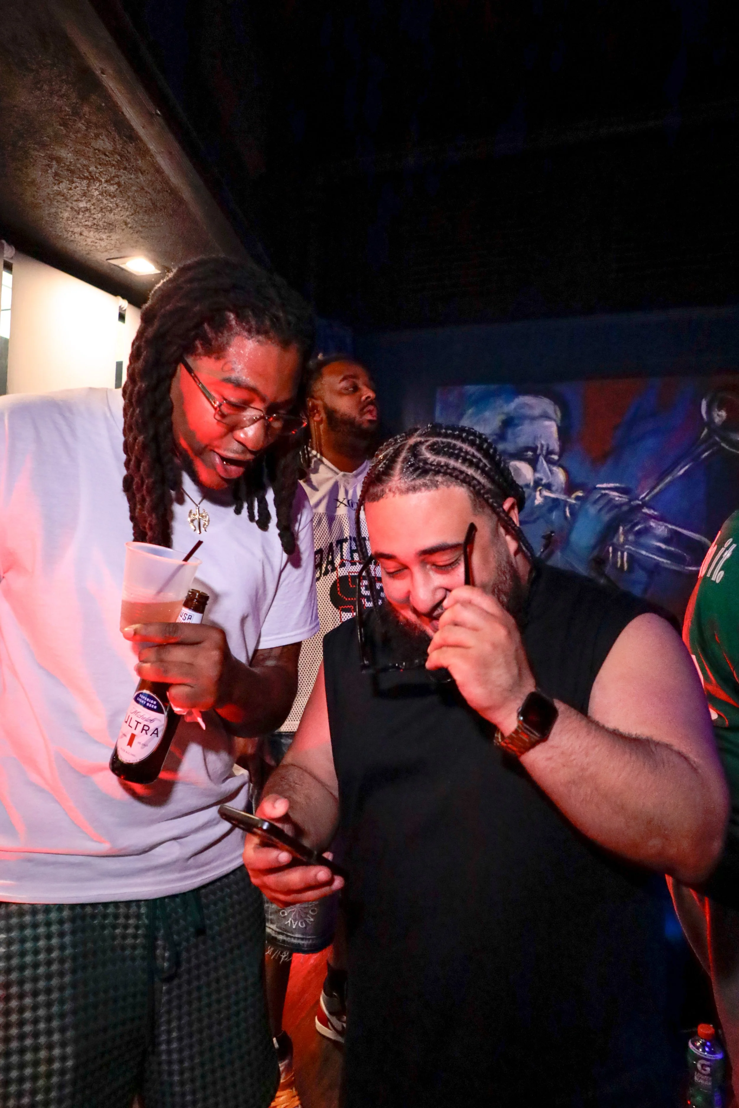 Three men in a dimly lit room looking at a phone, with colorful artwork on the wall behind them and drinks in hand.