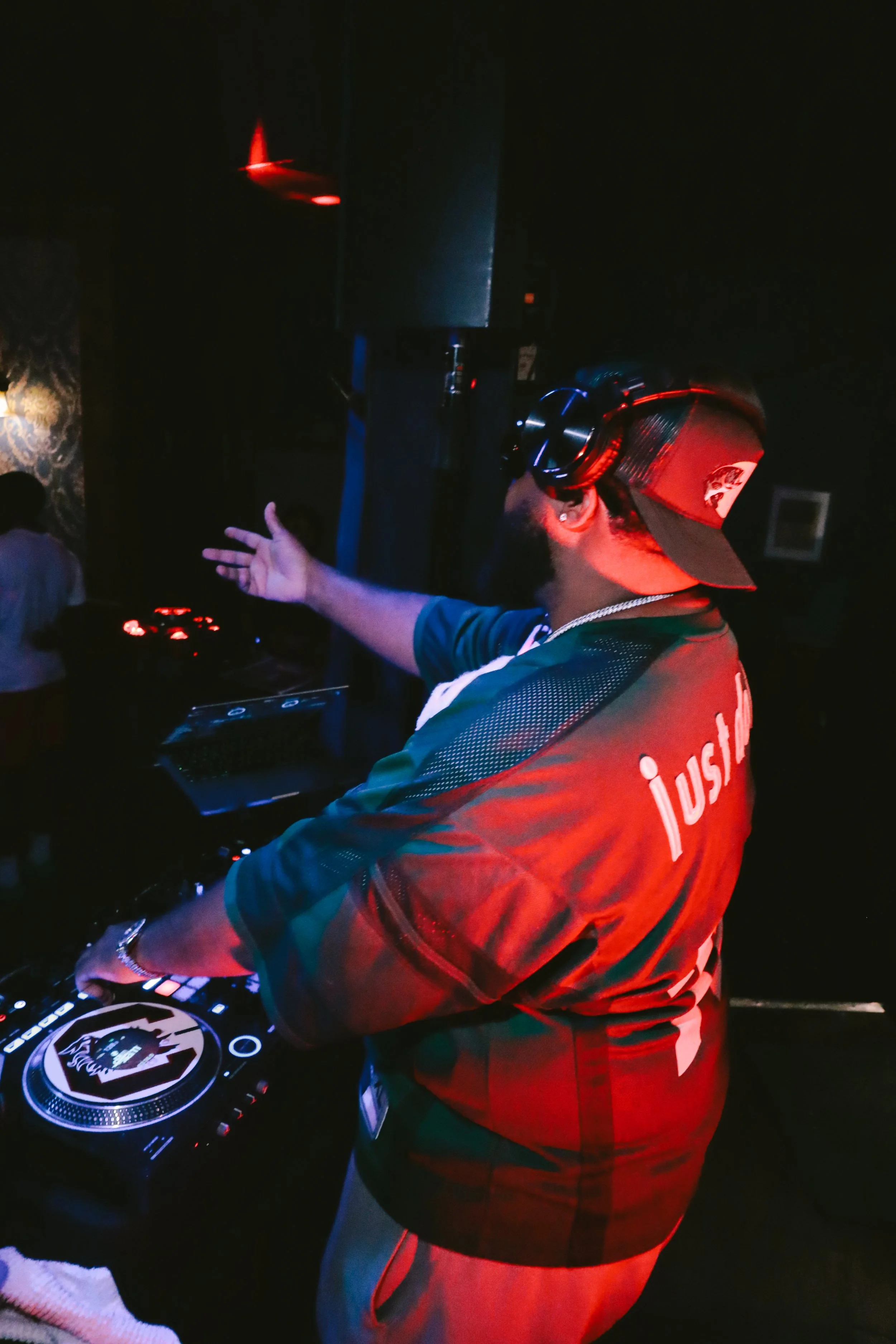 A DJ wearing a red baseball cap, black headphones, and a red jacket with 'JUSE' on the back, is performing at a nightclub or party, with a dark background and some people visible.