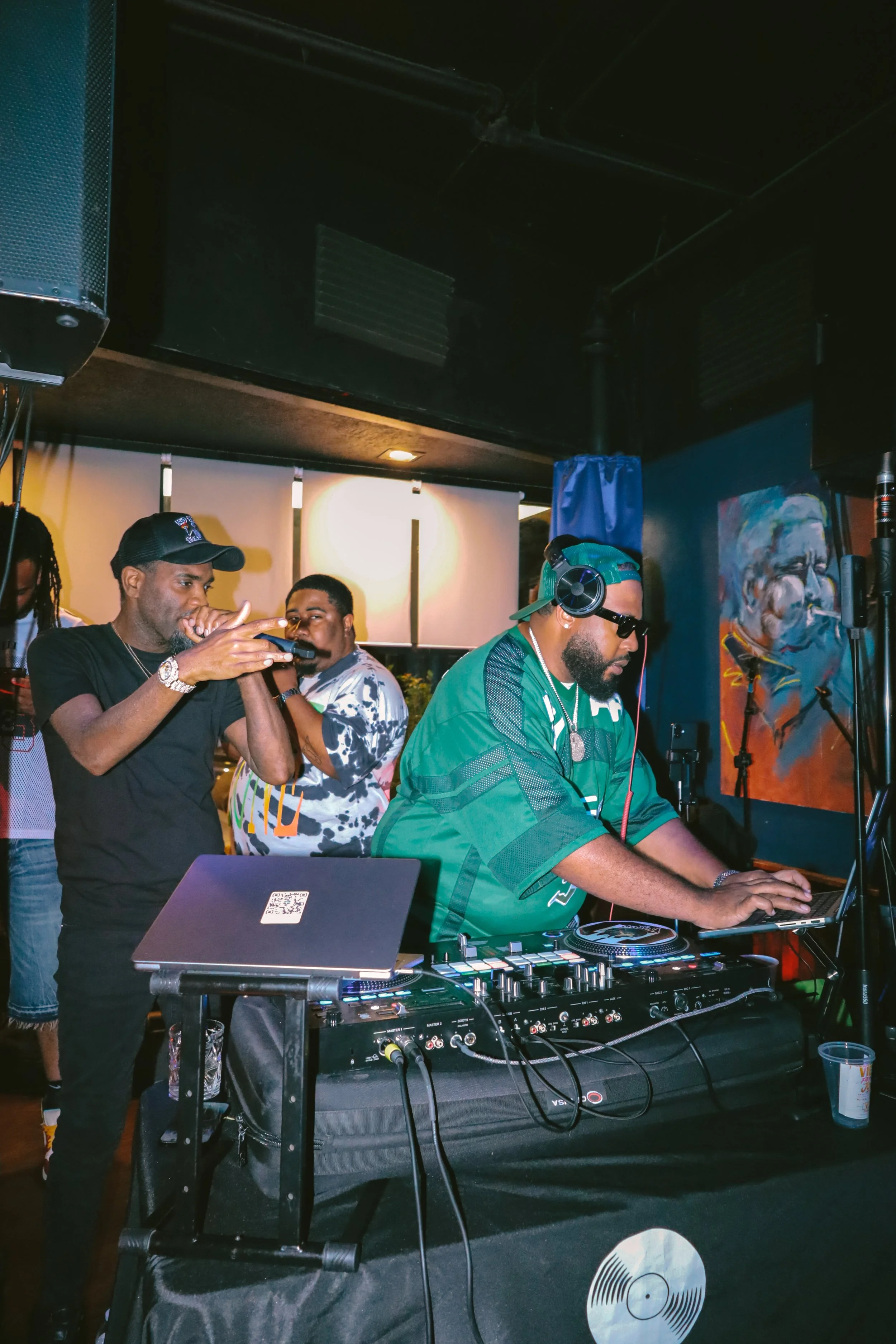 DJ iAM DJing at a party or event with DJ equipment in the foreground, and a colorful portrait painting on the wall in the background.