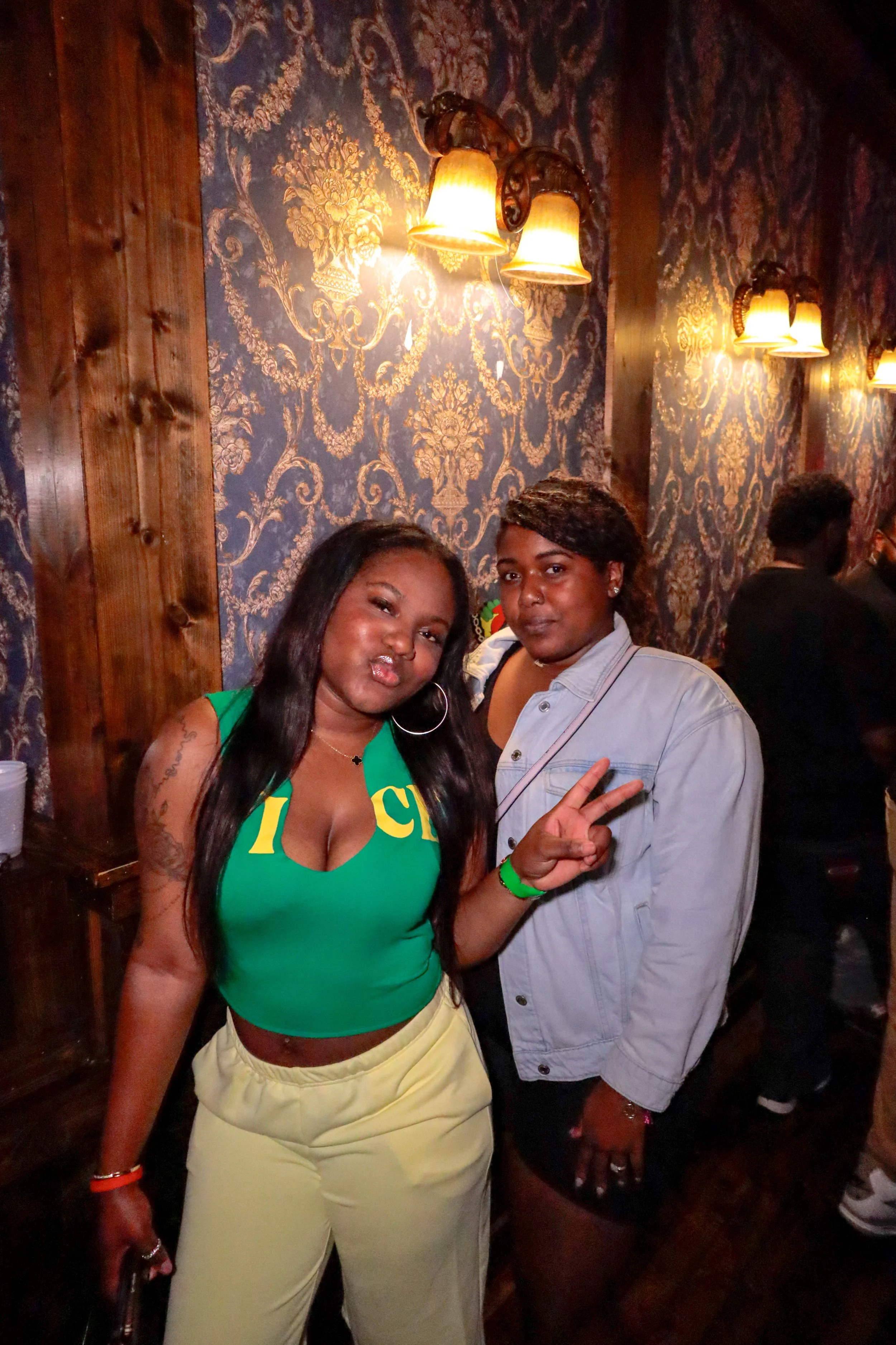 Two women posing together in a dimly lit room with ornate wallpaper and wall-mounted lamps. One woman is wearing a green top with Greek letters and yellow pants, making a peace sign. The other woman is wearing a light gray shirt. They are smiling and