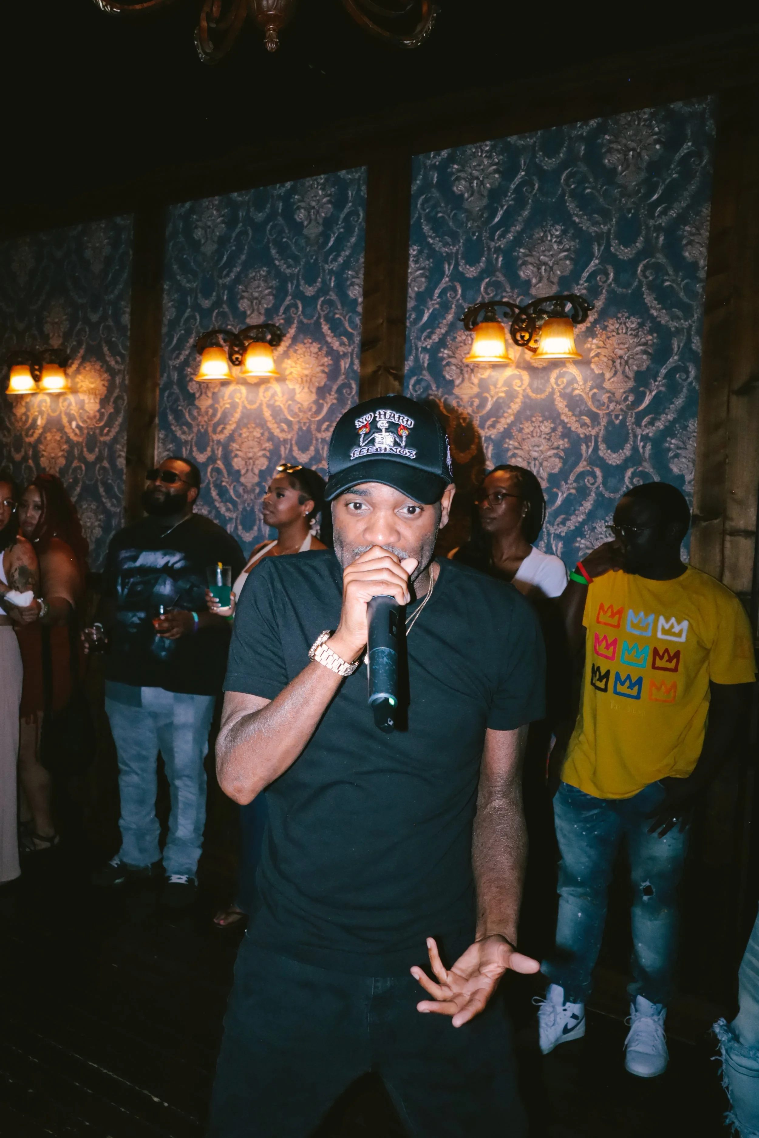 A man in a black t-shirt and a cap with the words 'No Hard Feelings' is holding a microphone and singing or speaking at a social event. Behind him, a group of diverse people are gathered, some holding drinks, in a dimly lit room with ornate blue wall