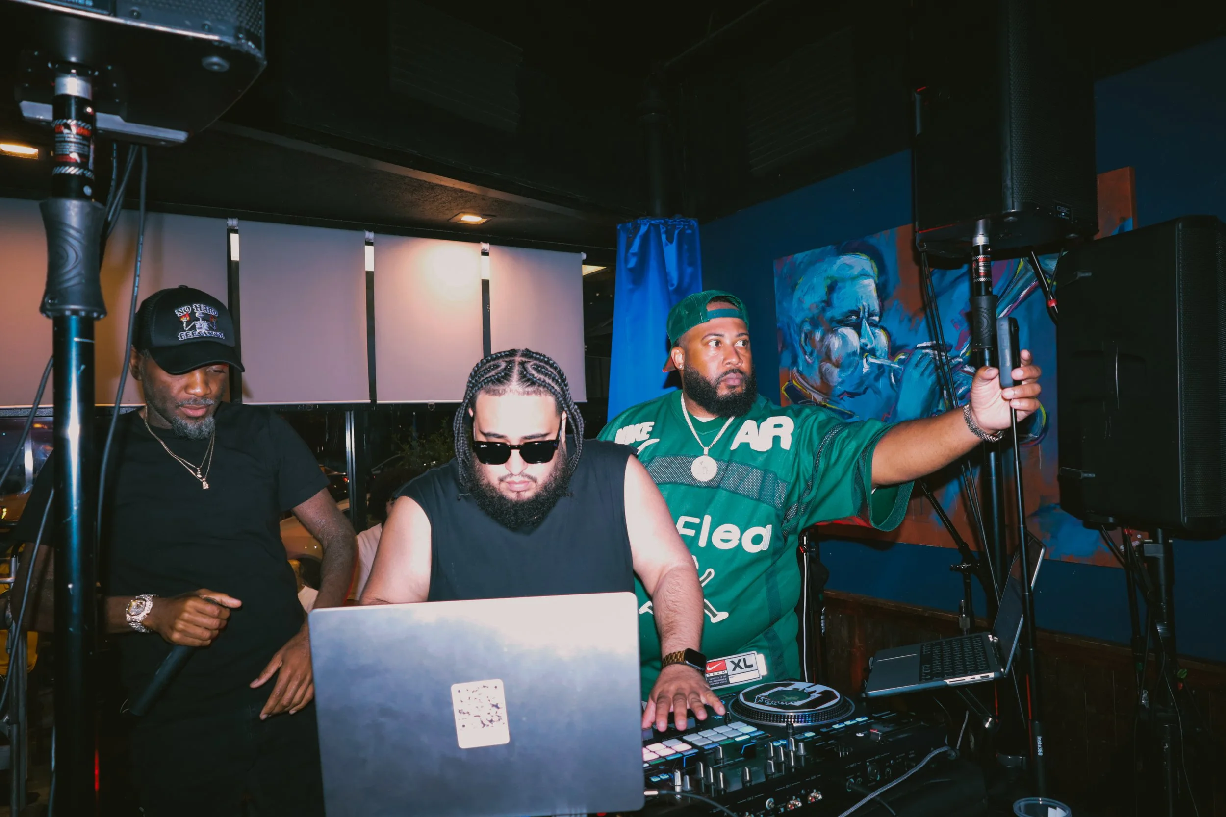 Three men, two with tattoos, one with braided hair, and another with a beard, are working with DJ equipment and a laptop at a party or event.
