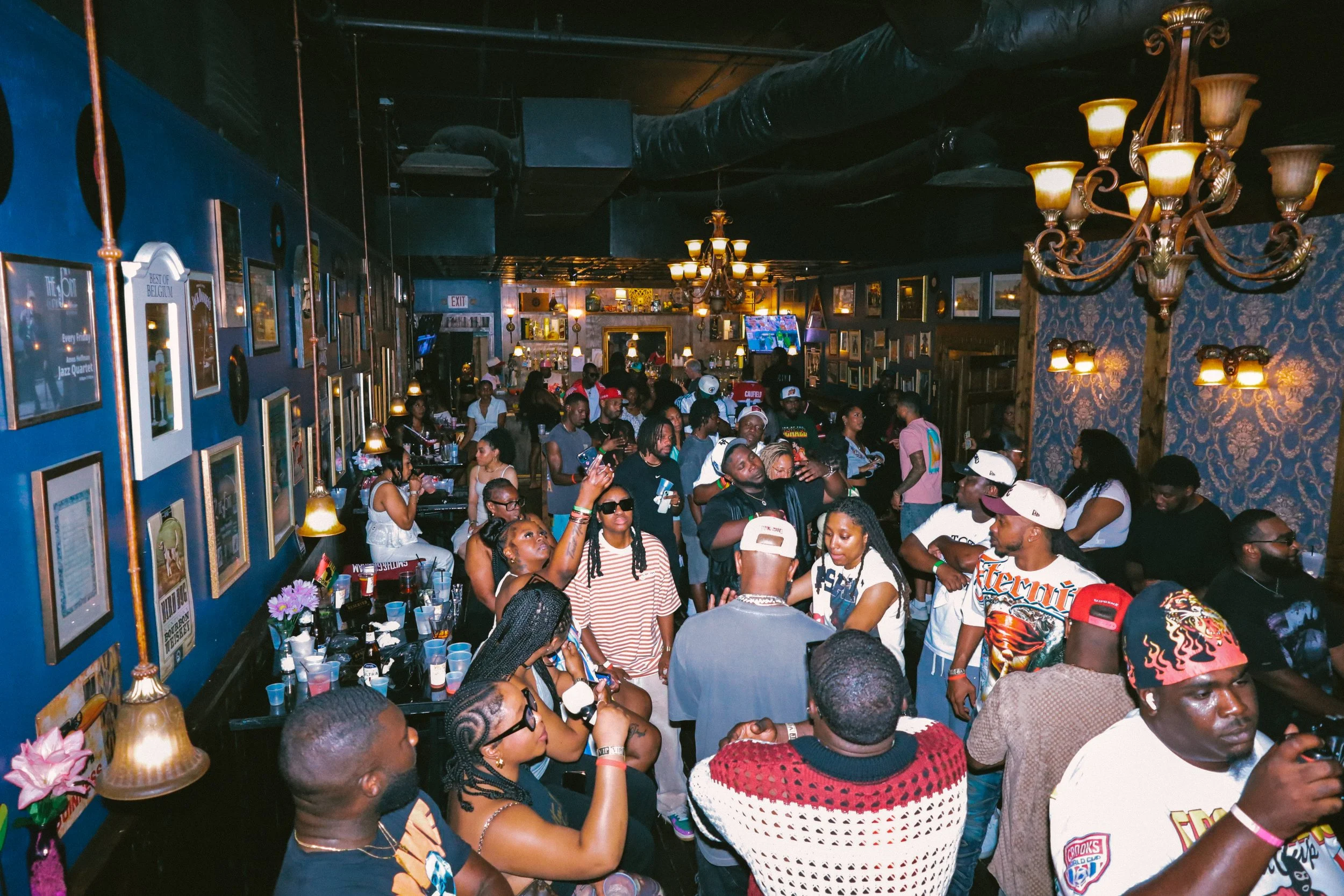 A lively indoor gathering with many people sitting and standing, socializing, in a decorated venue with framed pictures on the walls, chandeliers, and dim lighting.