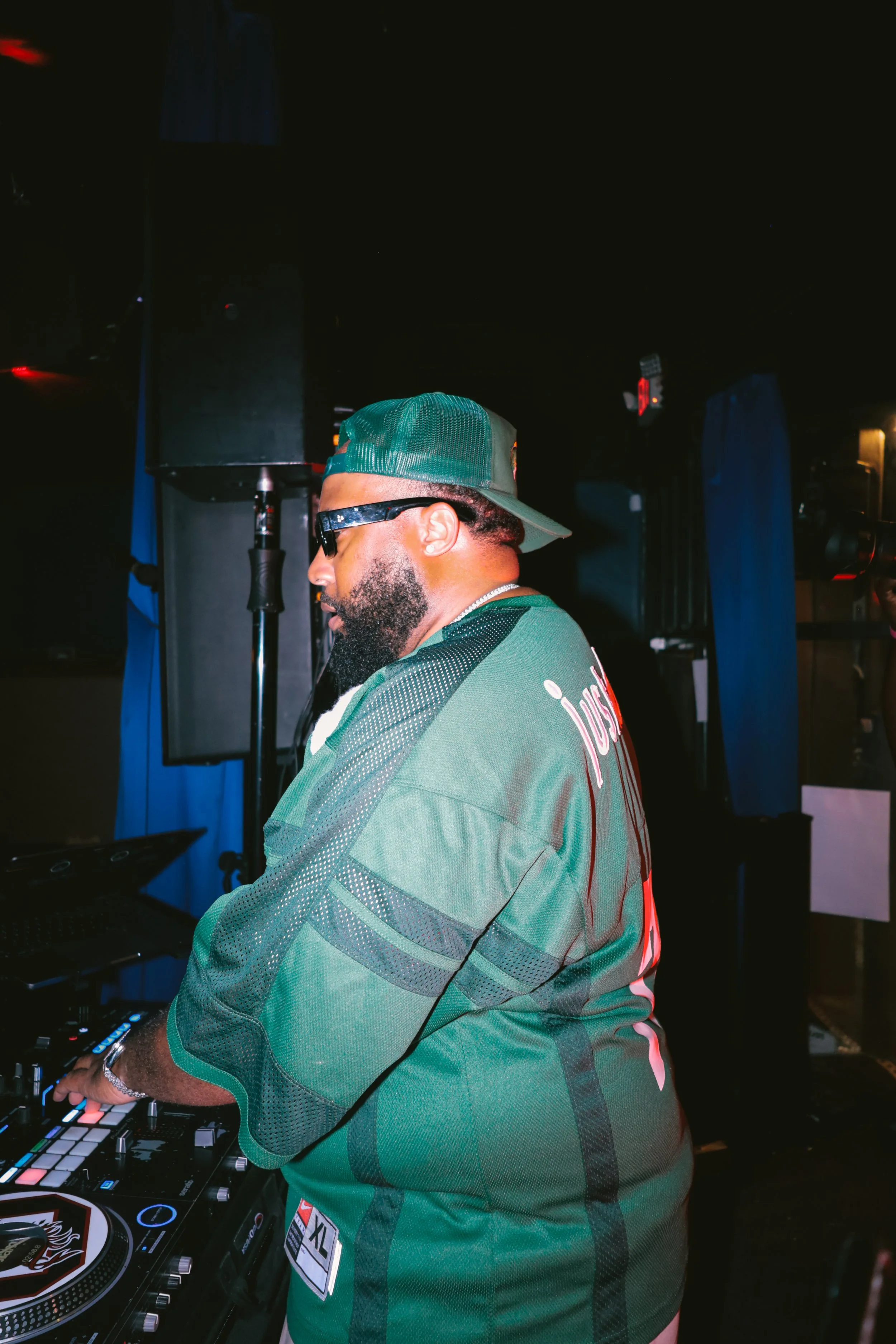 A DJ wearing sunglasses, a cap, and a green jersey with black accents is operating a DJ controller with multiple colorful pads in a dimly lit club or event space.