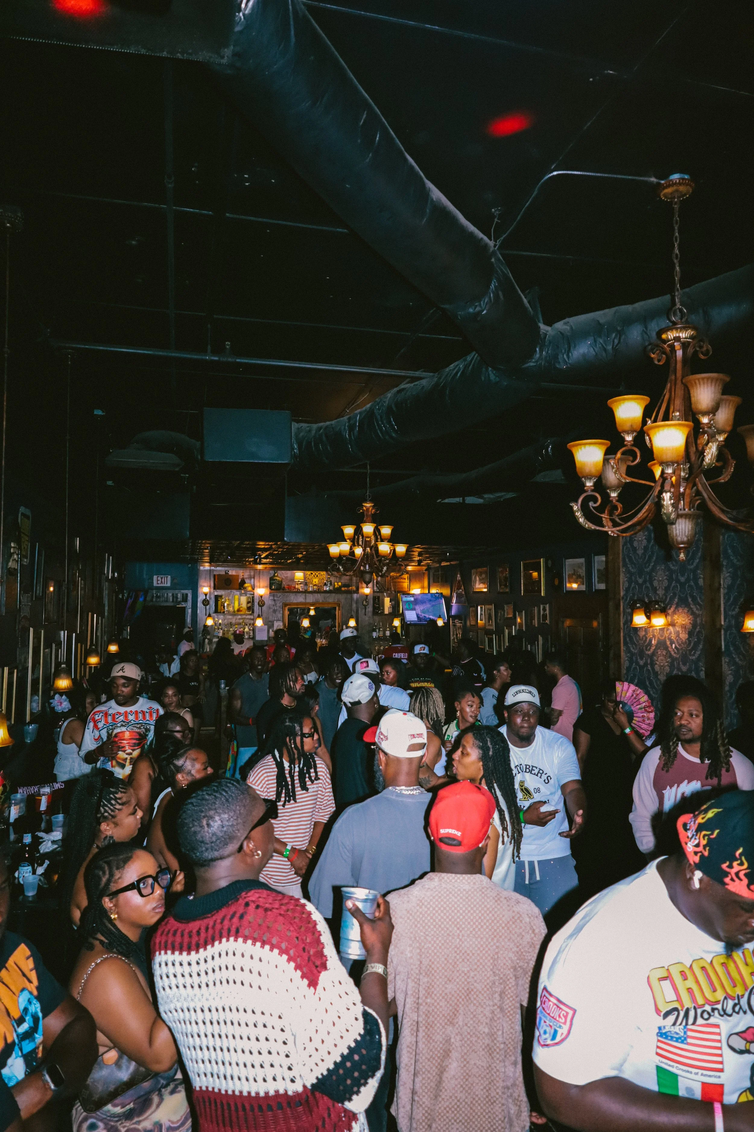 A crowded indoor event with many people standing close together, some holding drinks. There are chandeliers hanging from the ceiling, and the atmosphere appears lively and social.
