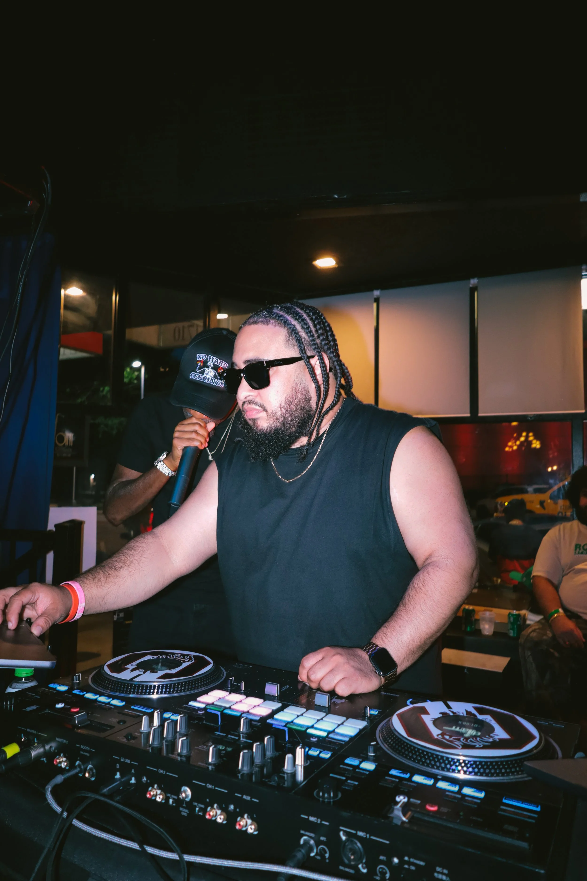 A man with braided hair, sunglasses, and a black sleeveless shirt is DJing on a mixer at a lively indoor event. Another person wearing a cap with the words 'No Fear' is holding a microphone behind him. There are tables with drinks and people in the b