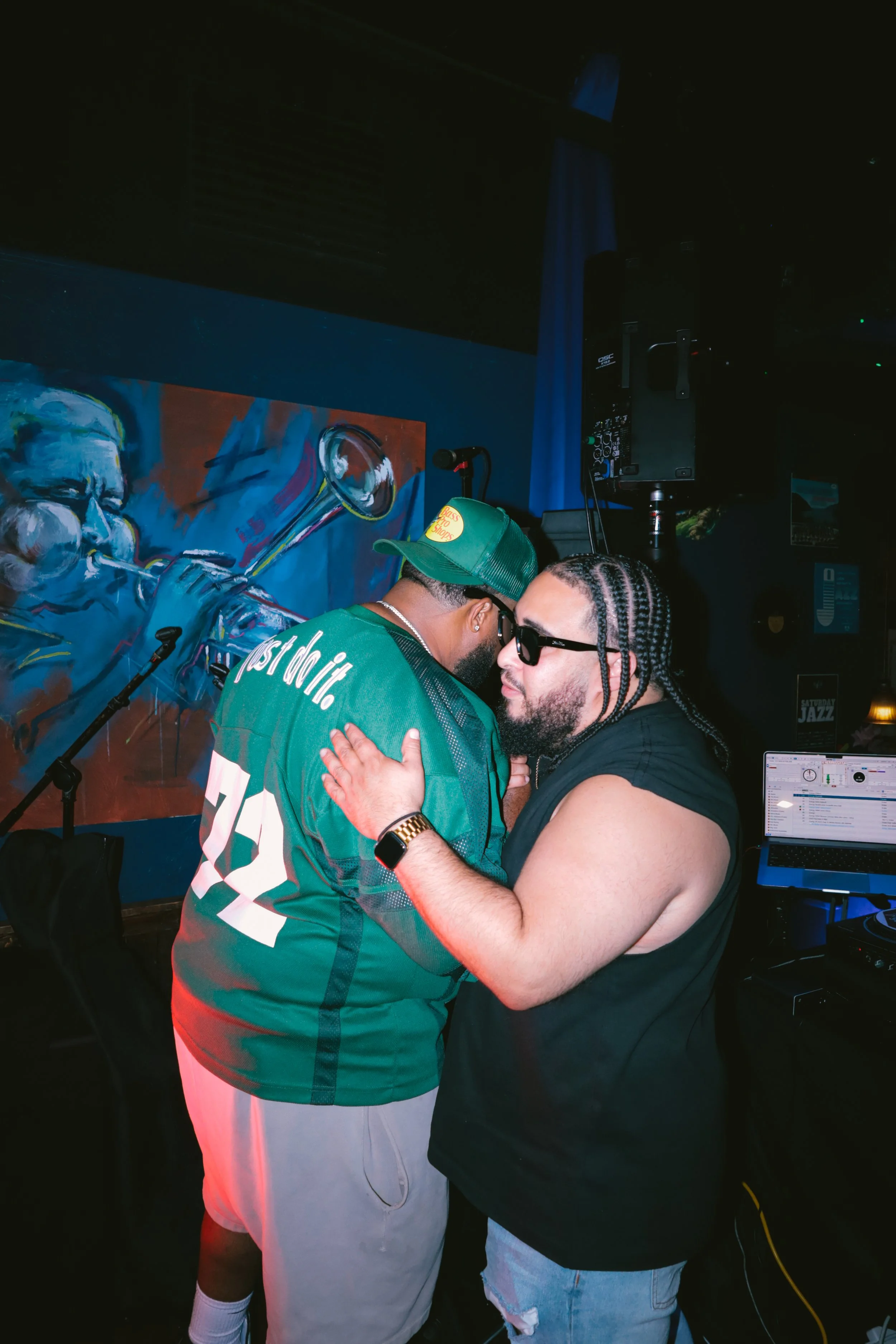 Two men are hugging in a club or music venue, with a colorful painting of a trumpet on the wall behind them, sound equipment, and a laptop in the background.