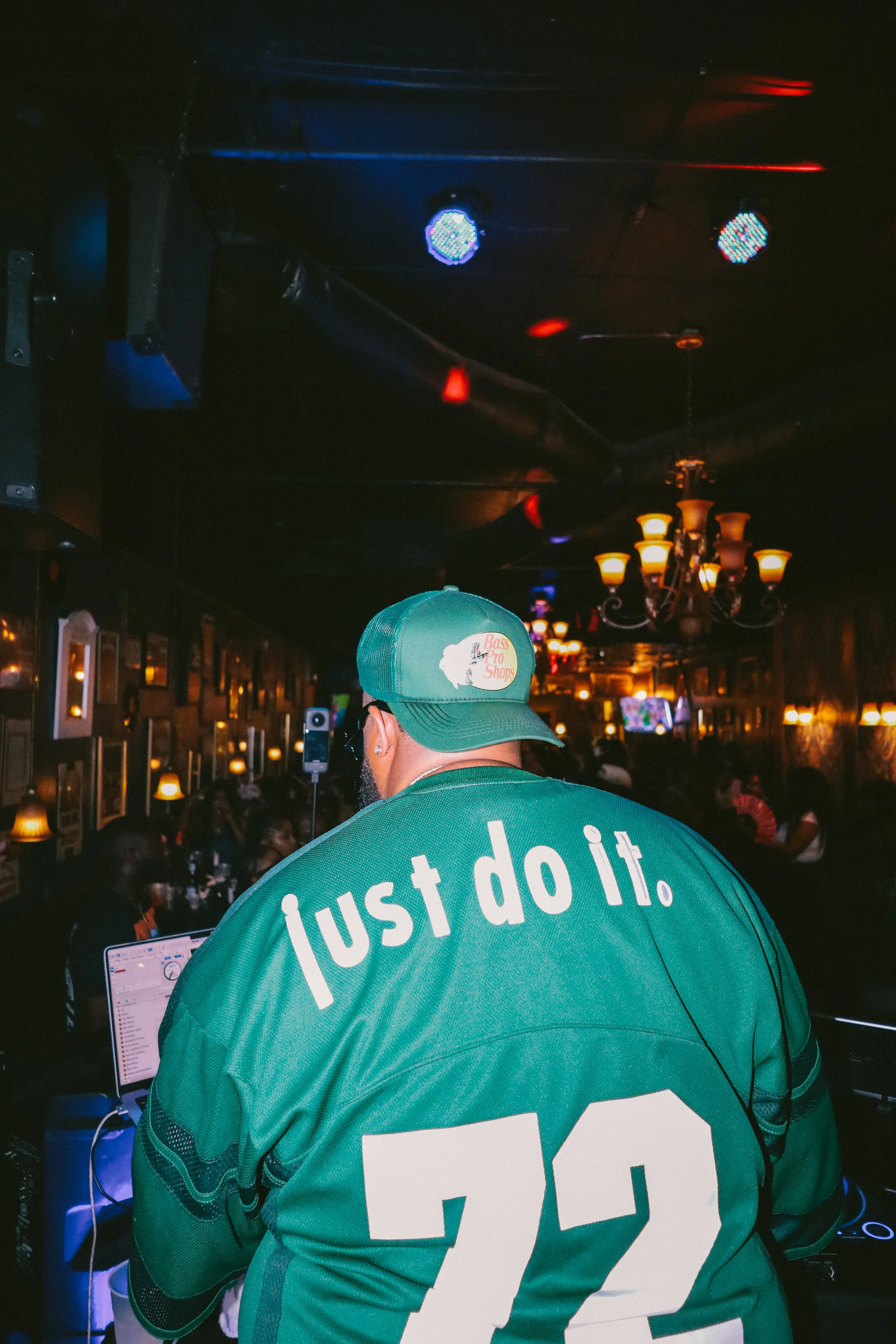 A person wearing a teal baseball jersey and cap with the Nike slogan 'Just Do It' is in a dimly lit bar or club, with a chandelier and other patrons in the background, engrossed in a laptop screen.