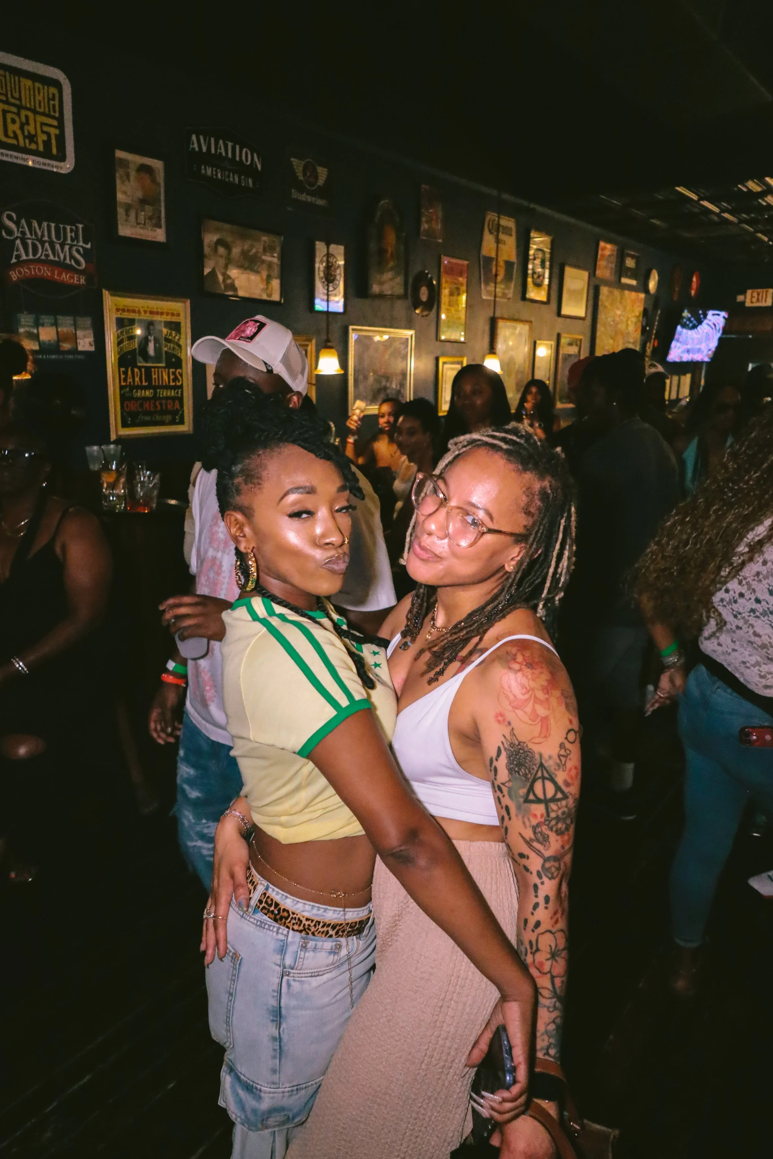 Two women embracing and smiling in a dimly lit bar or club with people and framed pictures on the wall in the background.