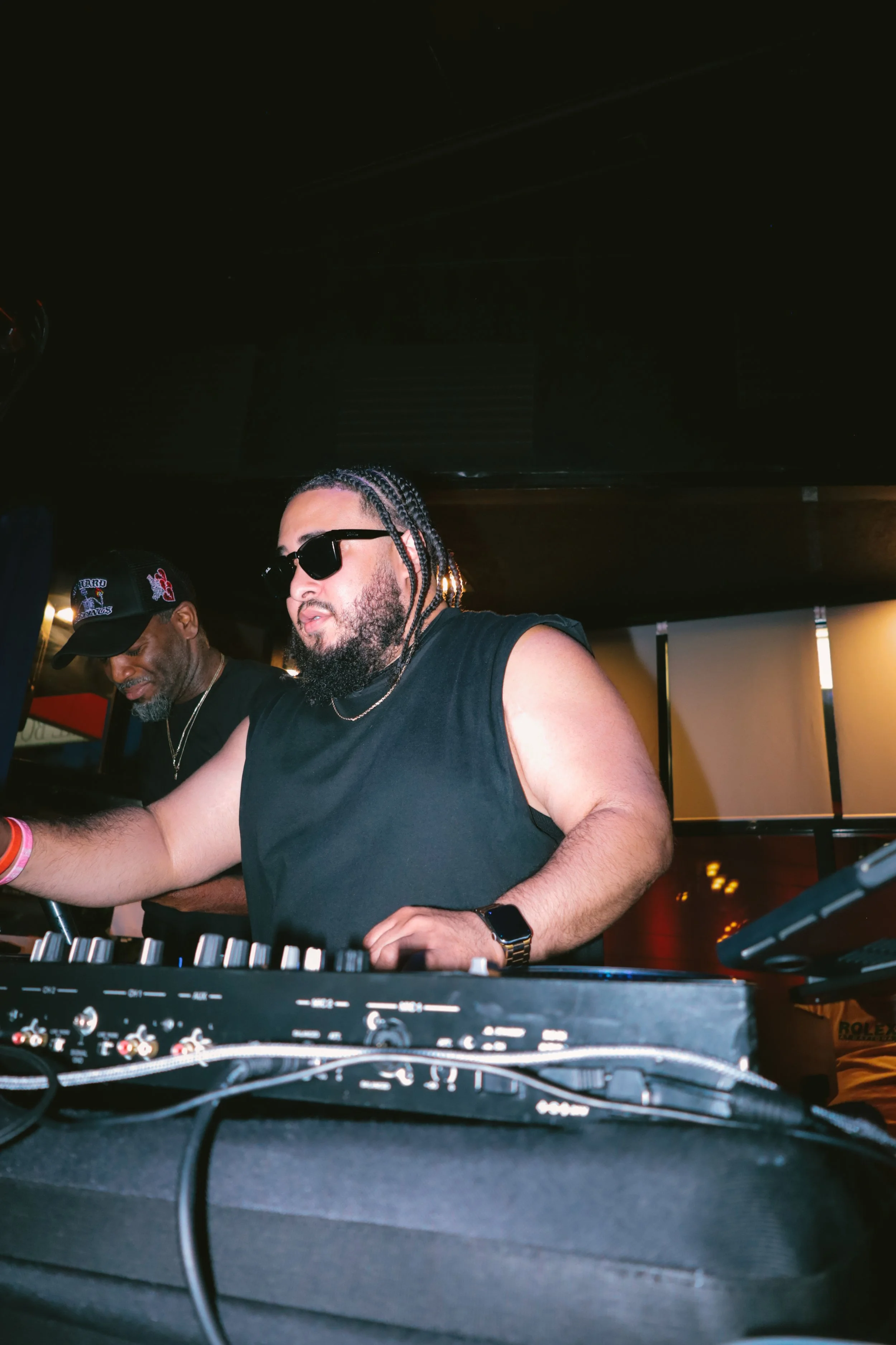 A DJ with braided hair, sunglasses, a beard, wearing a black sleeveless shirt, and a smartwatch, performing at a nightclub or music event. Another man wearing a cap is next to him, and DJ equipment is in the foreground.
