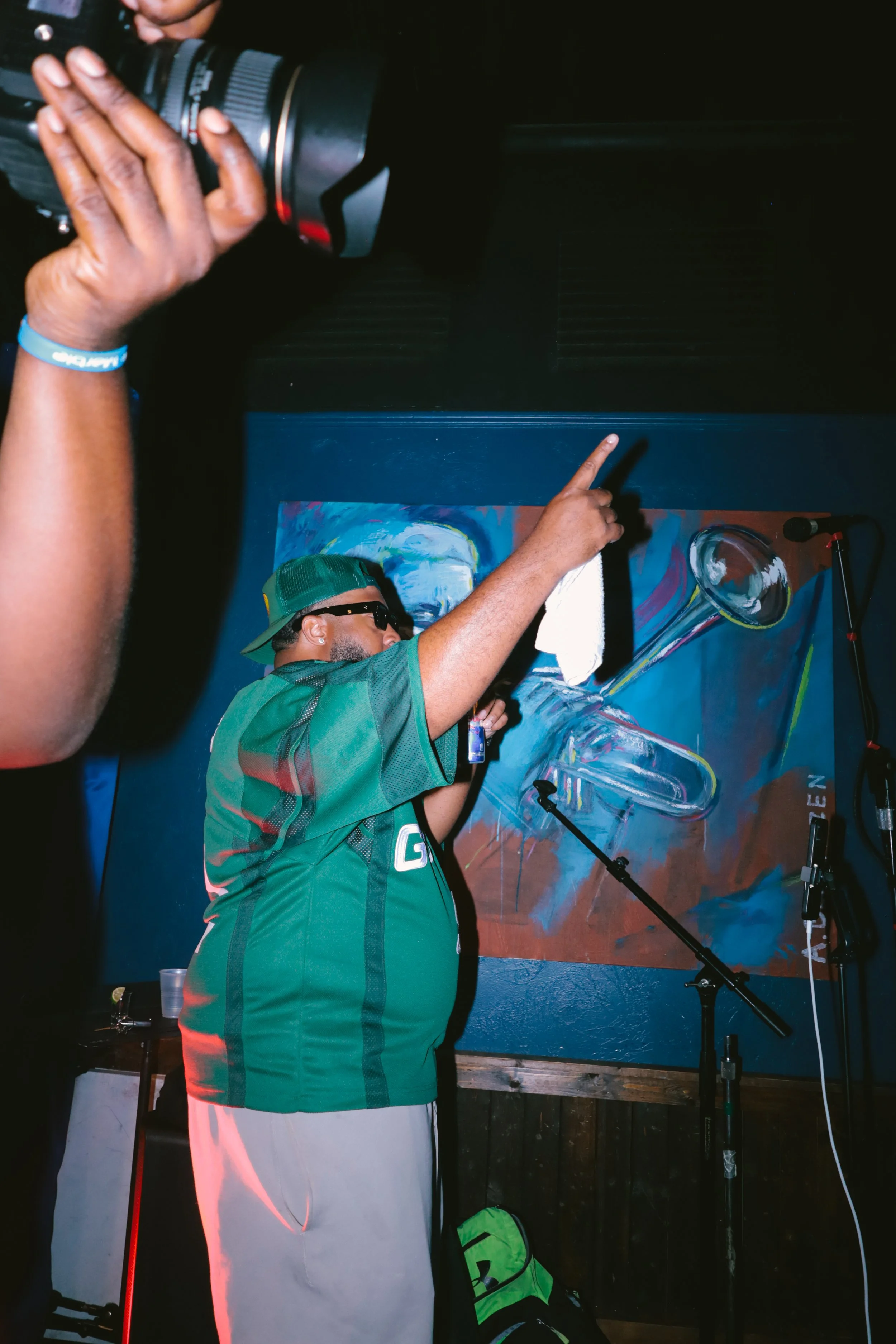 DJ iAM wearing a green cap, sunglasses, and a green jersey is on stage, pointing upward with his right hand while holding a microphone in his left hand. There is a colorful abstract painting behind him and a microphone stand in front of him.