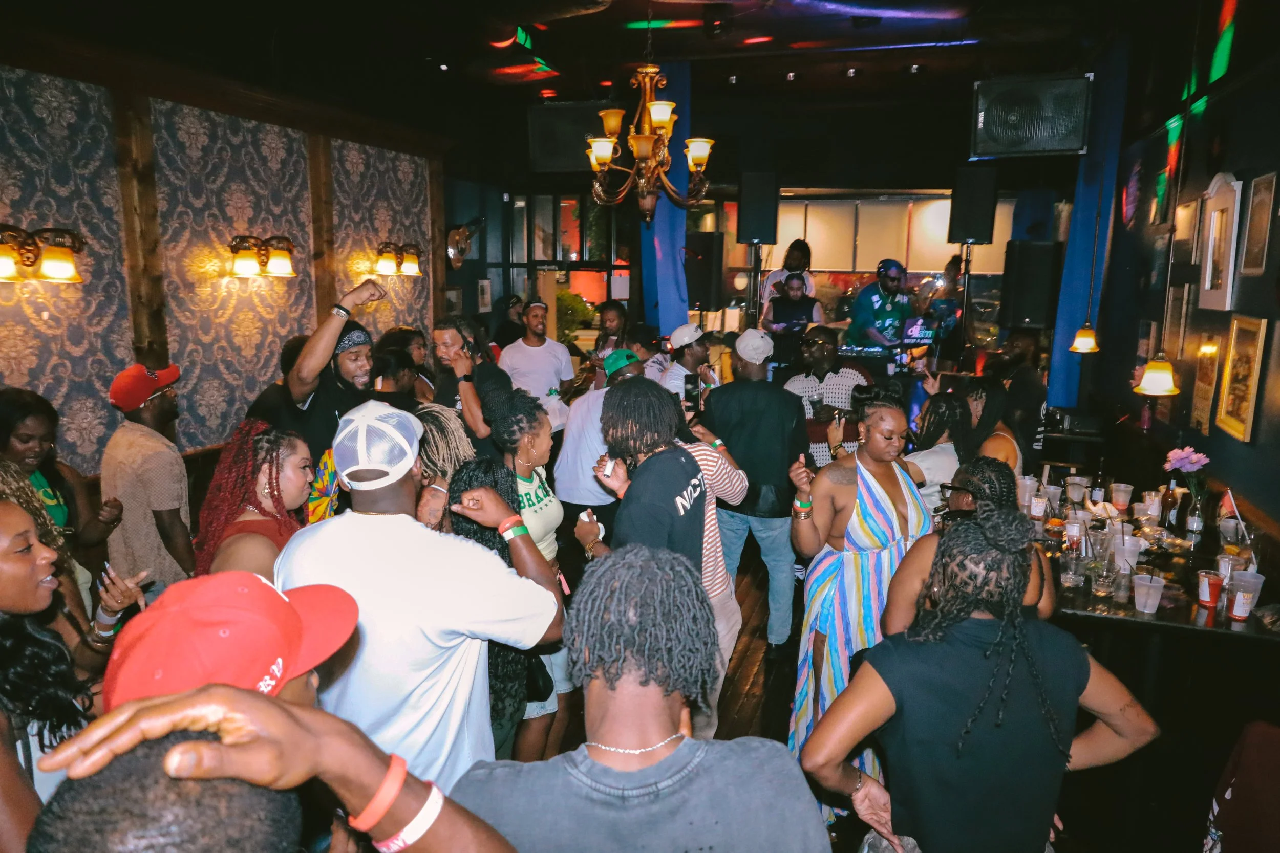 A lively indoor party scene with many people dancing and socializing. The room features dark walls with ornate wallpapers, warm lighting, and a DJ booth with equipment. People are wearing casual and colorful clothes, some with hats and accessories. A
