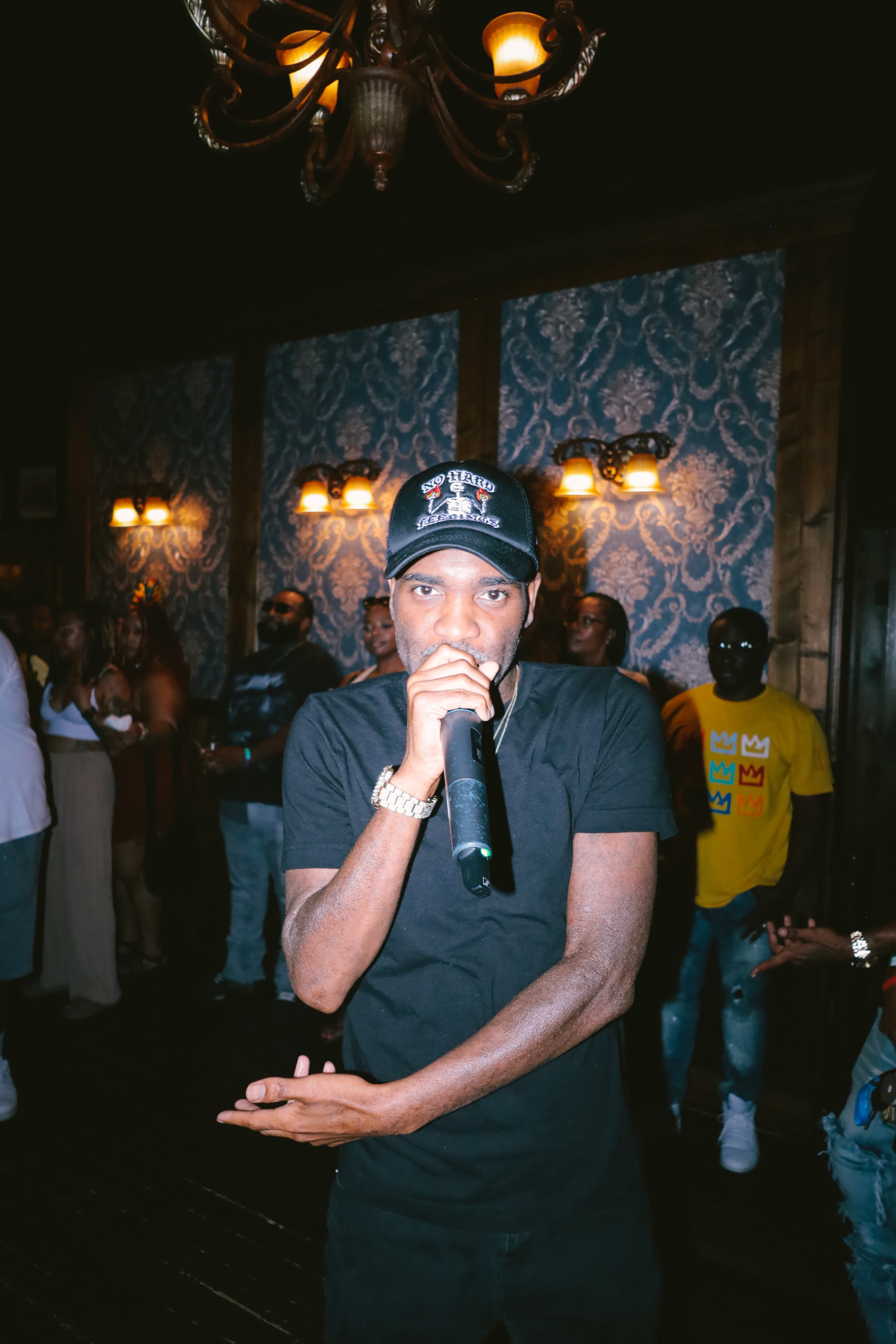A man wearing a black t-shirt, a black cap, and a watch, holding a microphone in a dimly lit room with a chandelier and patterned wallpaper, surrounded by a group of people.