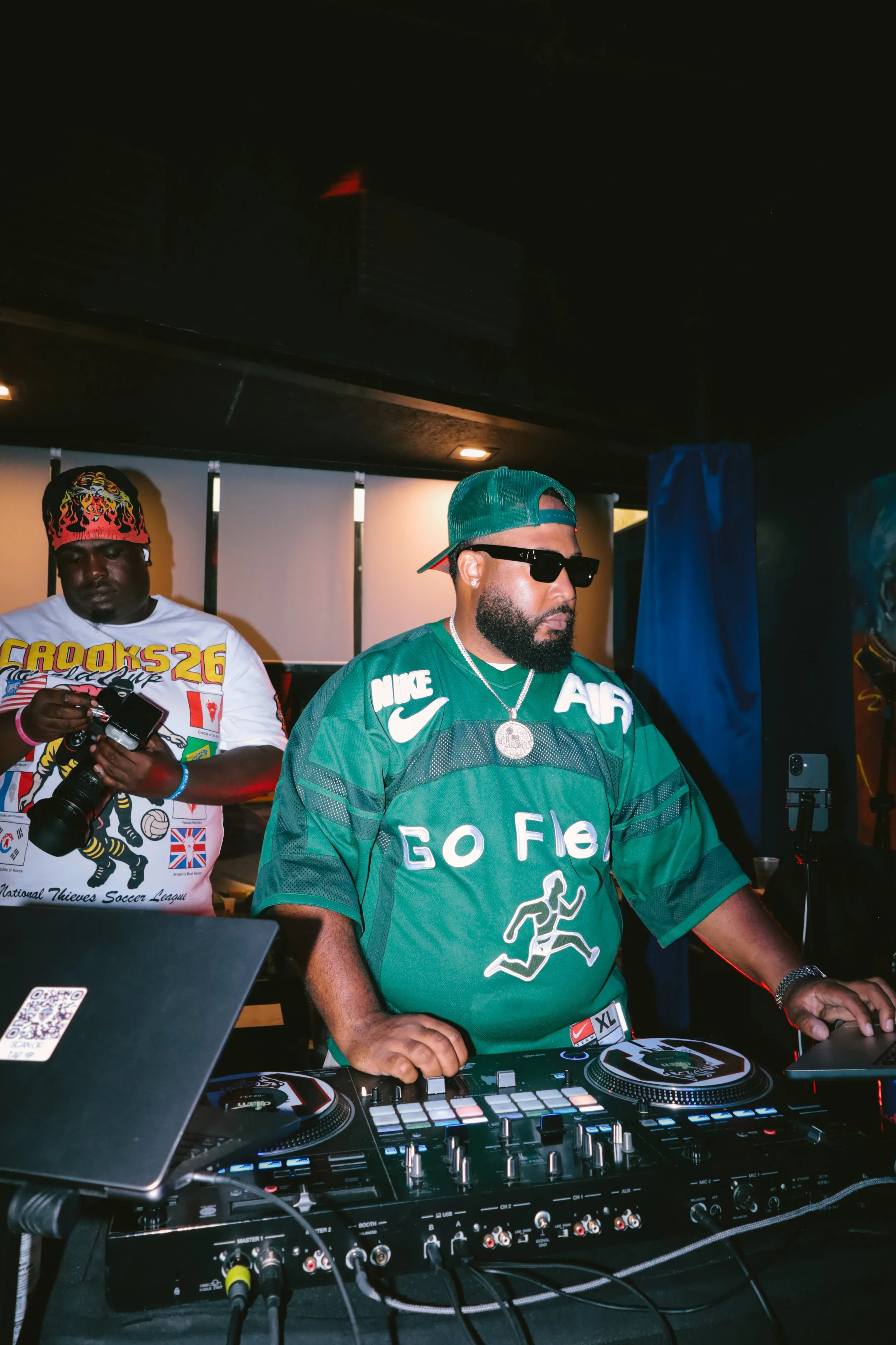 Two men are at a DJ setup with equipment including turntables, a mixer, and a laptop. The man in the foreground is wearing a green sports jersey, a green cap, and sunglasses, while the man in the background is wearing a white T-shirt with a soccer le