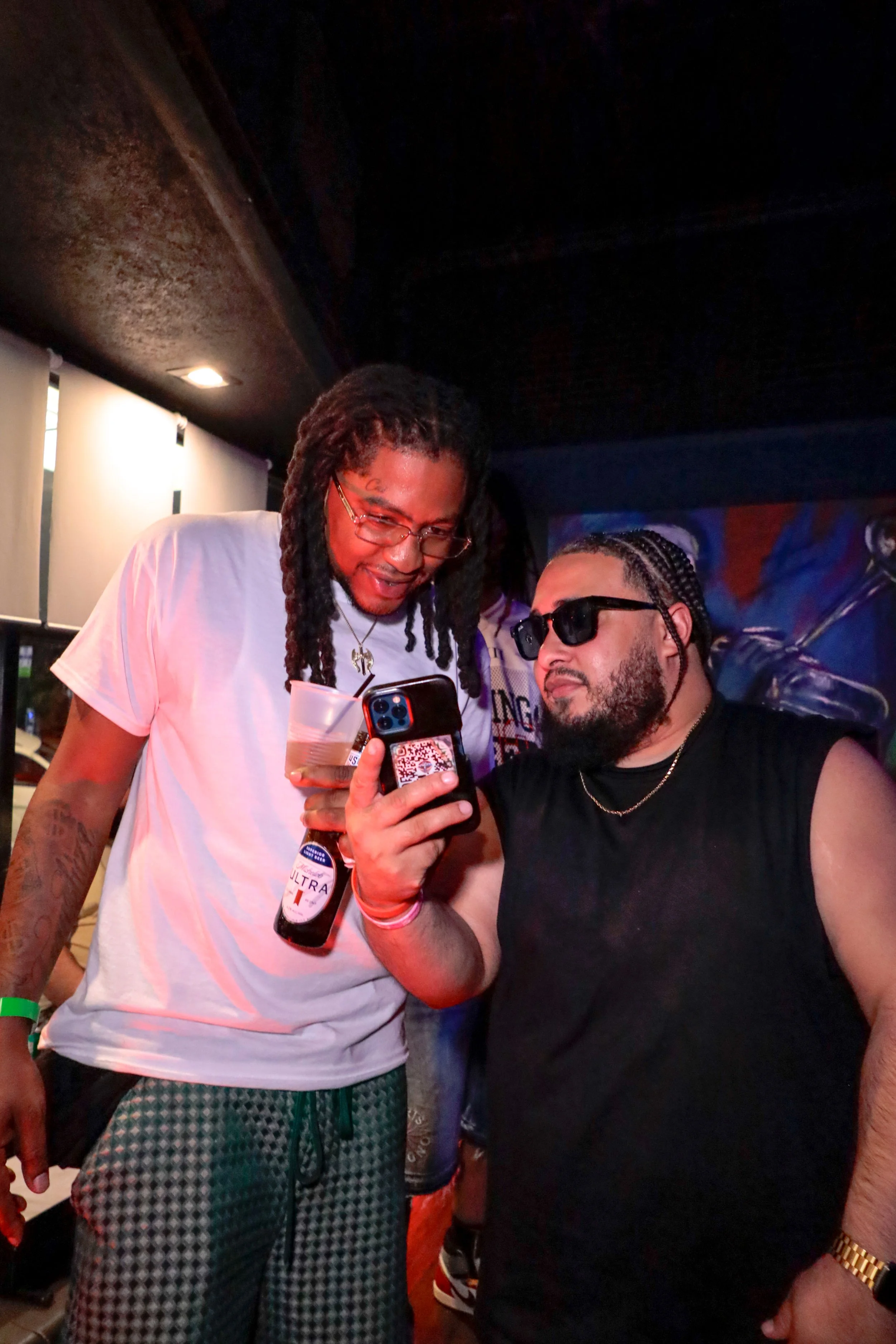 Two men are looking at a phone together in a dimly lit indoor setting. One man has dreadlocks, glasses, and is holding a drink and a phone with a QR code. The other man has braided hair, sunglasses, and is wearing a black sleeveless shirt and a gold 