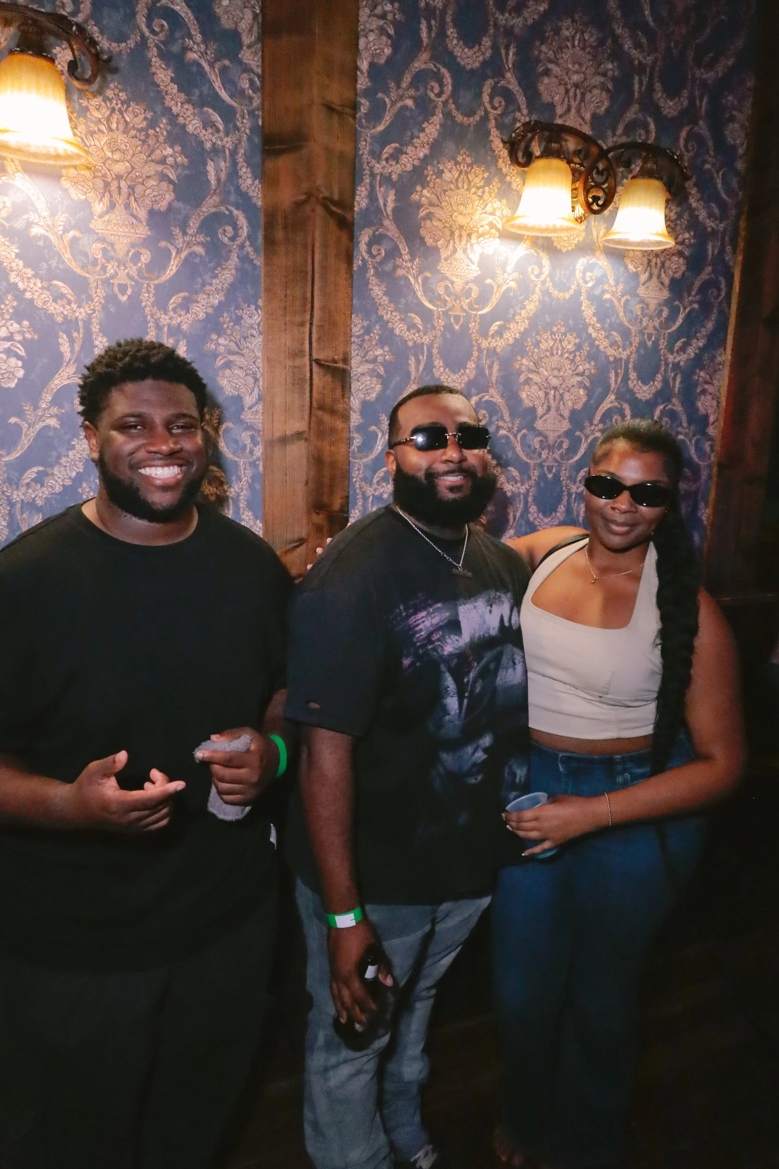Three people standing together in a dimly lit room with wallpapered walls and wall-mounted lamps, smiling for a photo. Two men and one woman, wearing casual clothing and sunglasses.
