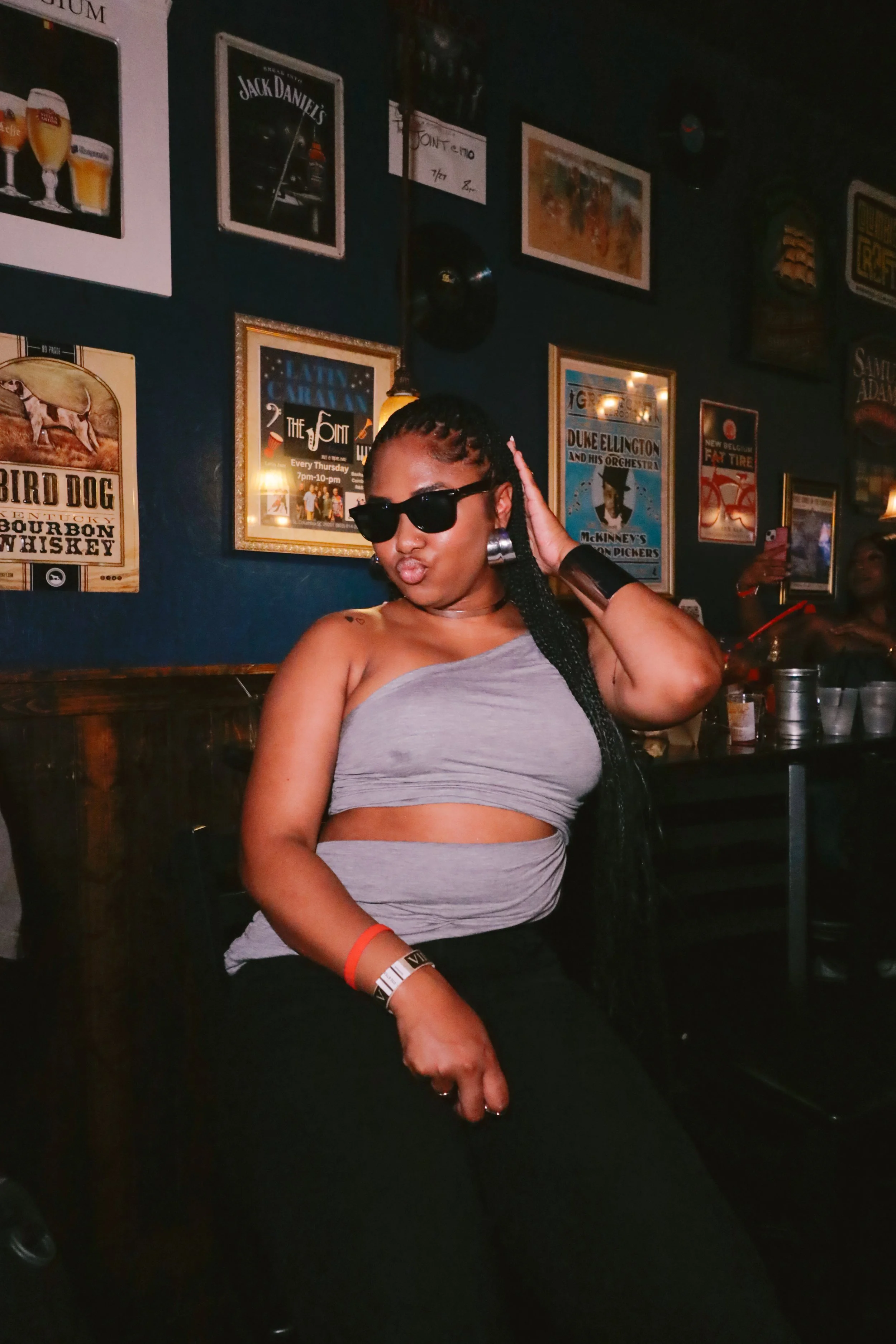 A woman wearing sunglasses, a gray crop top, and black pants posing in a dimly lit bar or restaurant with framed posters on the wall behind her.