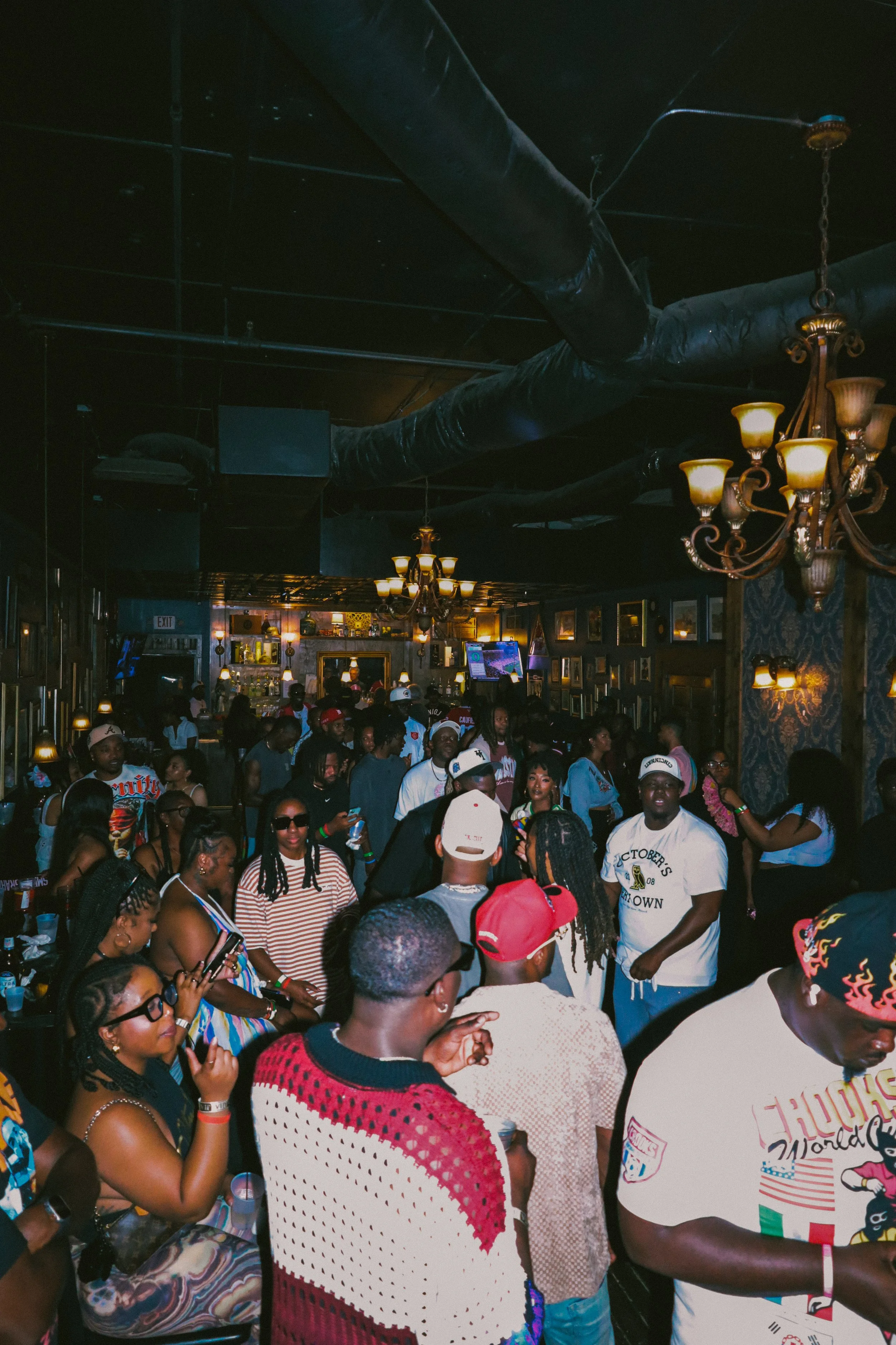 Crowd of people gathered inside a dimly lit bar or restaurant, with dark walls, ornate chandeliers, and framed pictures hanging on the walls.