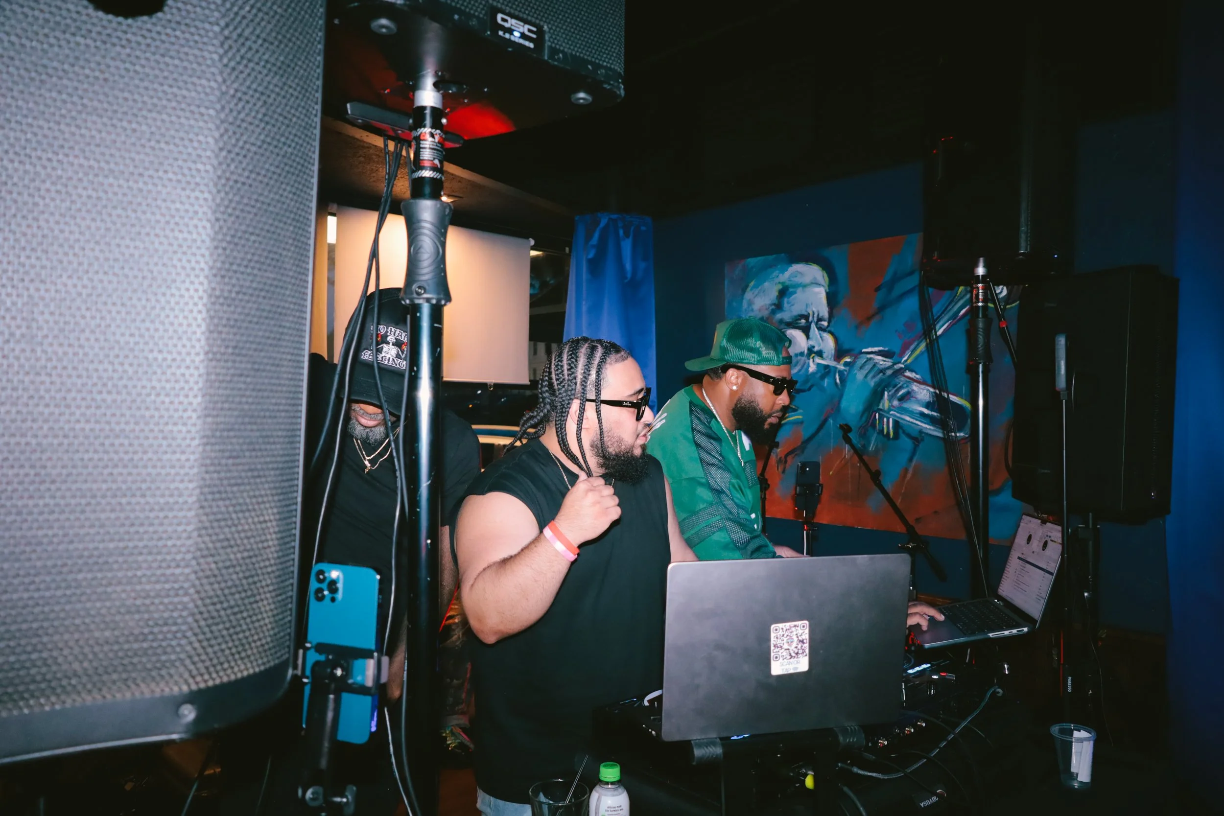 Three men in a dimly lit room, working on laptops, with sound equipment and a colorful abstract mural in the background.