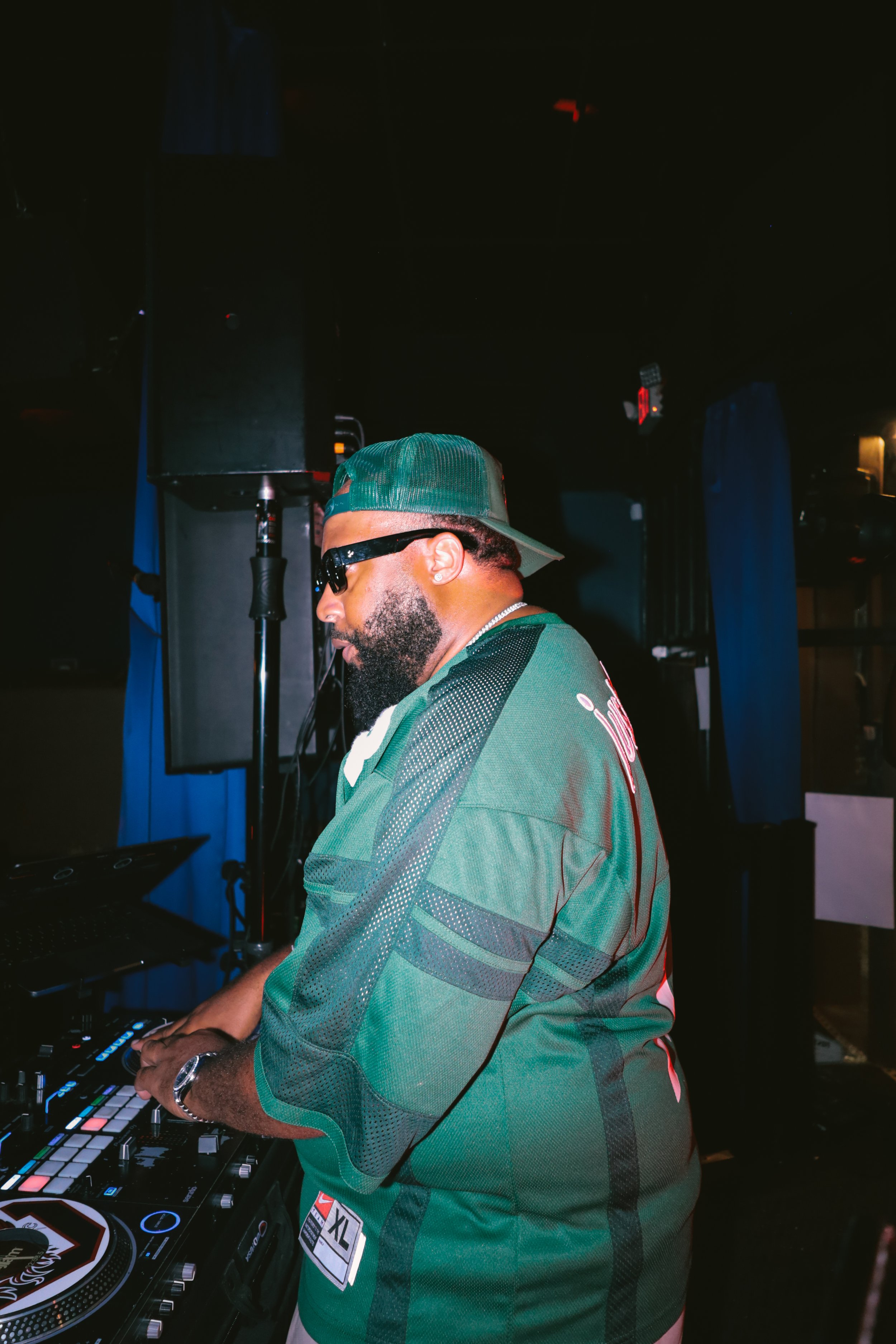 A DJ wearing a green baseball cap, sunglasses, and a green sports jersey, operating DJ equipment in a dark club setting.