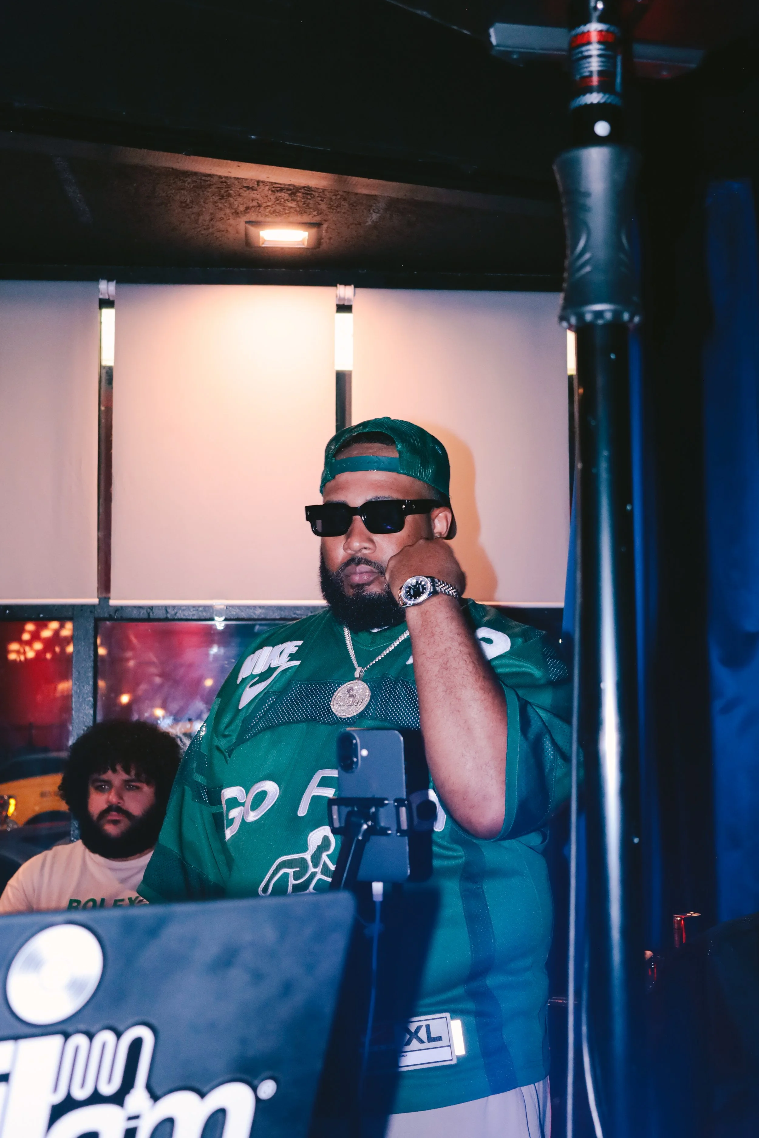 A man wearing sunglasses, a green sports jersey, a wristwatch, and a green cap backwards, is seen with his hand on his face, standing in front of a mirror. There is another man with a beard and curly hair in the background. The room appears to be a c