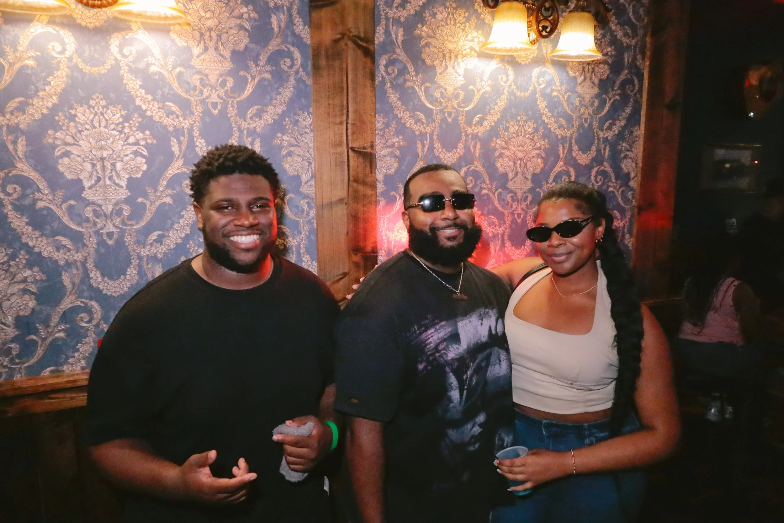 Three people standing together in front of a floral wallpapered wall with warm lighting, smiling, two wearing sunglasses and one holding a drink.
