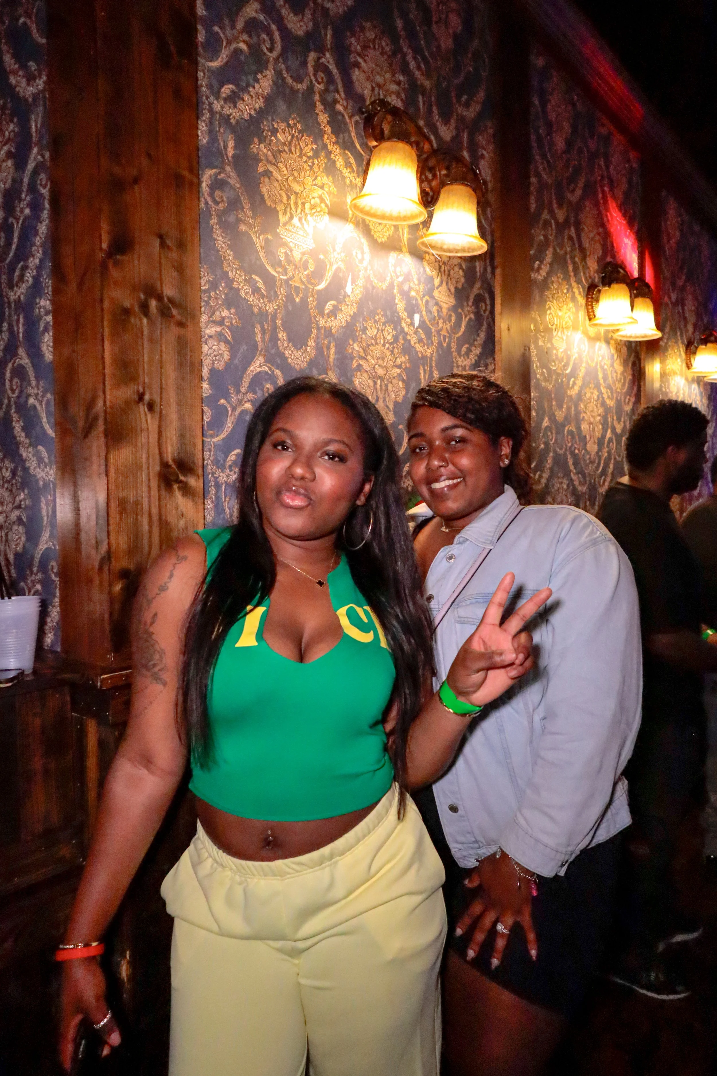 Two women posing for a photo in a dimly lit venue with ornate wallpaper and wall-mounted lamps. One woman is wearing a green crop top with yellow accents and yellow pants, making a peace sign. The other woman is wearing a light-colored button-up shir