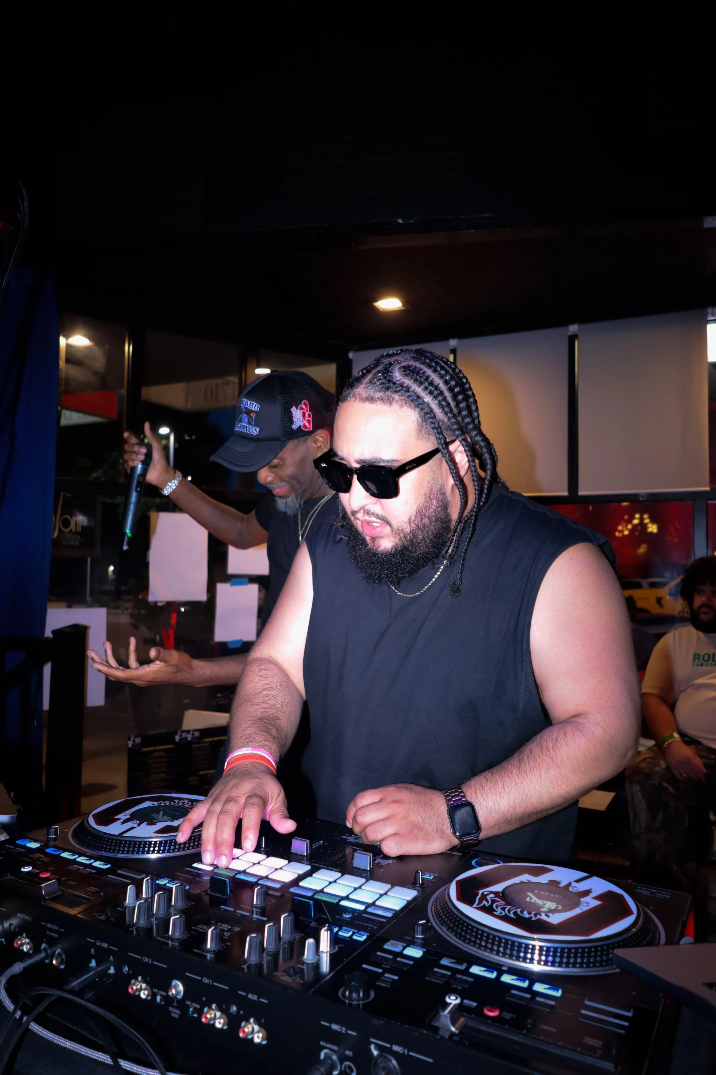 Two men are DJing at a crowded indoor event. The man in the foreground wears sunglasses, a black sleeveless shirt, and has braided hair. He is operating DJ equipment, looking down at the mixer. The man in the background, wearing a black cap with patc