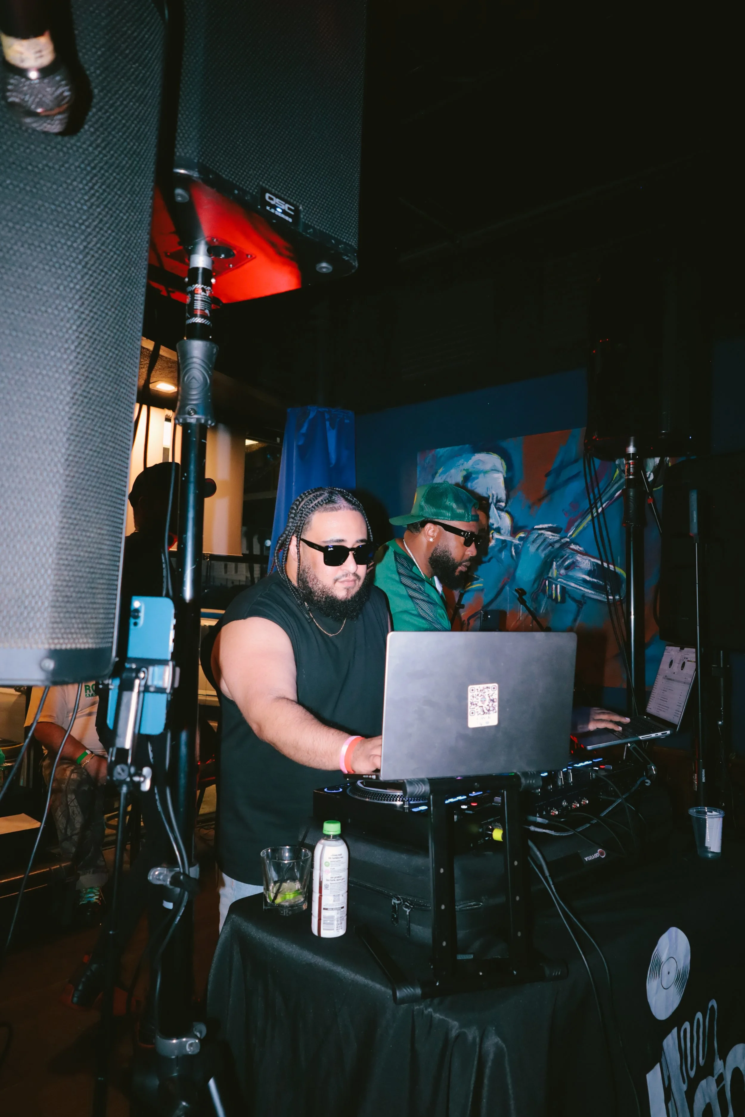 Two men are DJing at a party or event indoors, with one man wearing sunglasses and a black t-shirt, and the other wearing a green cap and sunglasses. There are speakers, laptops, and DJ equipment on a table, with a colorful mural painting in the back
