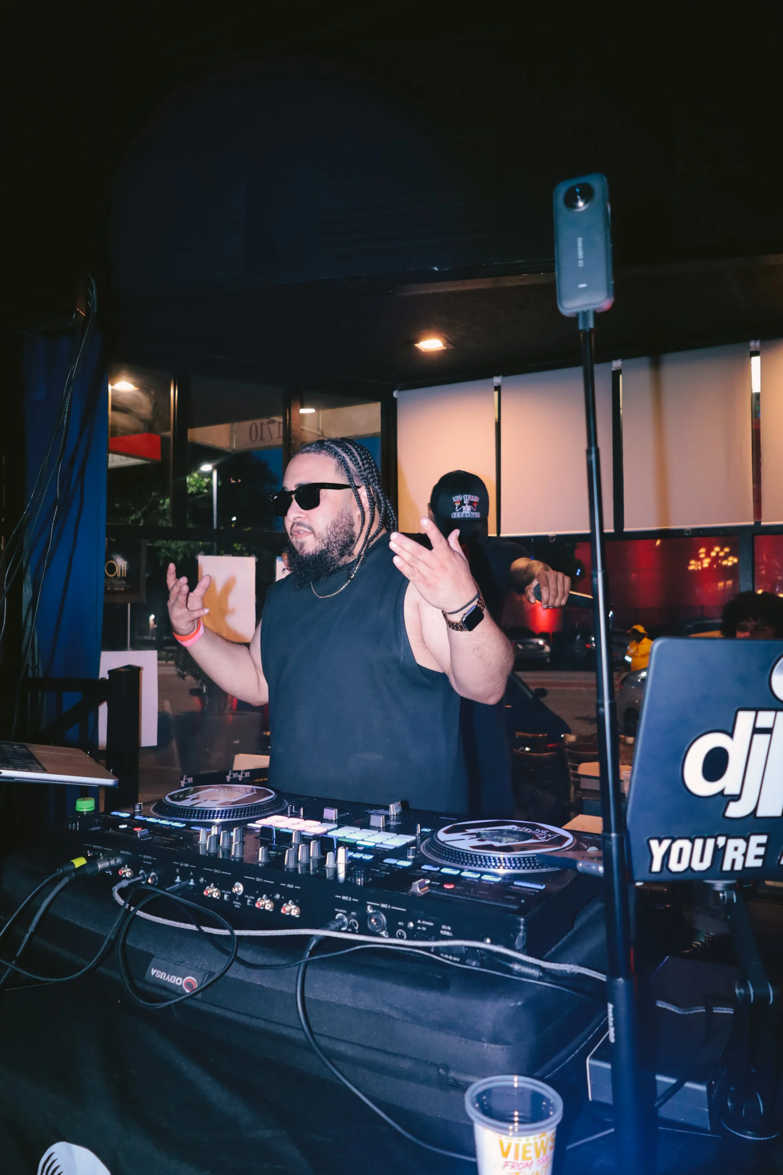 A DJ wearing sunglasses and a black sleeveless shirt, with braided hair, dancing or gesturing with his hands in a nightclub or music venue. The DJ setup is visible with turntables, mixer, and laptop. There is a crowd outside through the windows and a