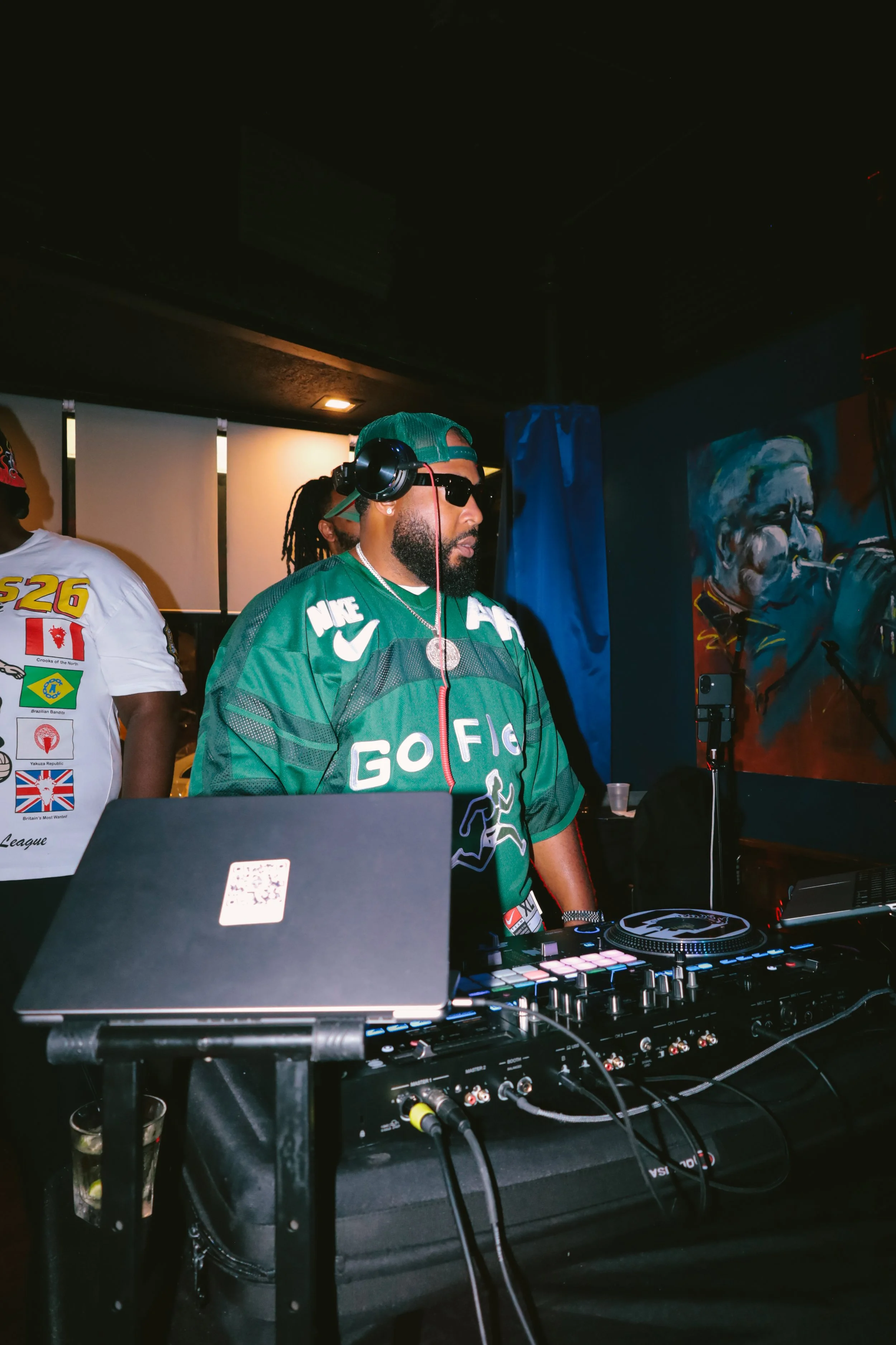 A DJ wearing sunglasses, a green cap, and a green shirt with white lettering is mixing music at a dark indoor event. The DJ is standing behind a professional DJ controller and laptop, with cables connected. There is a colorful painting of a person in