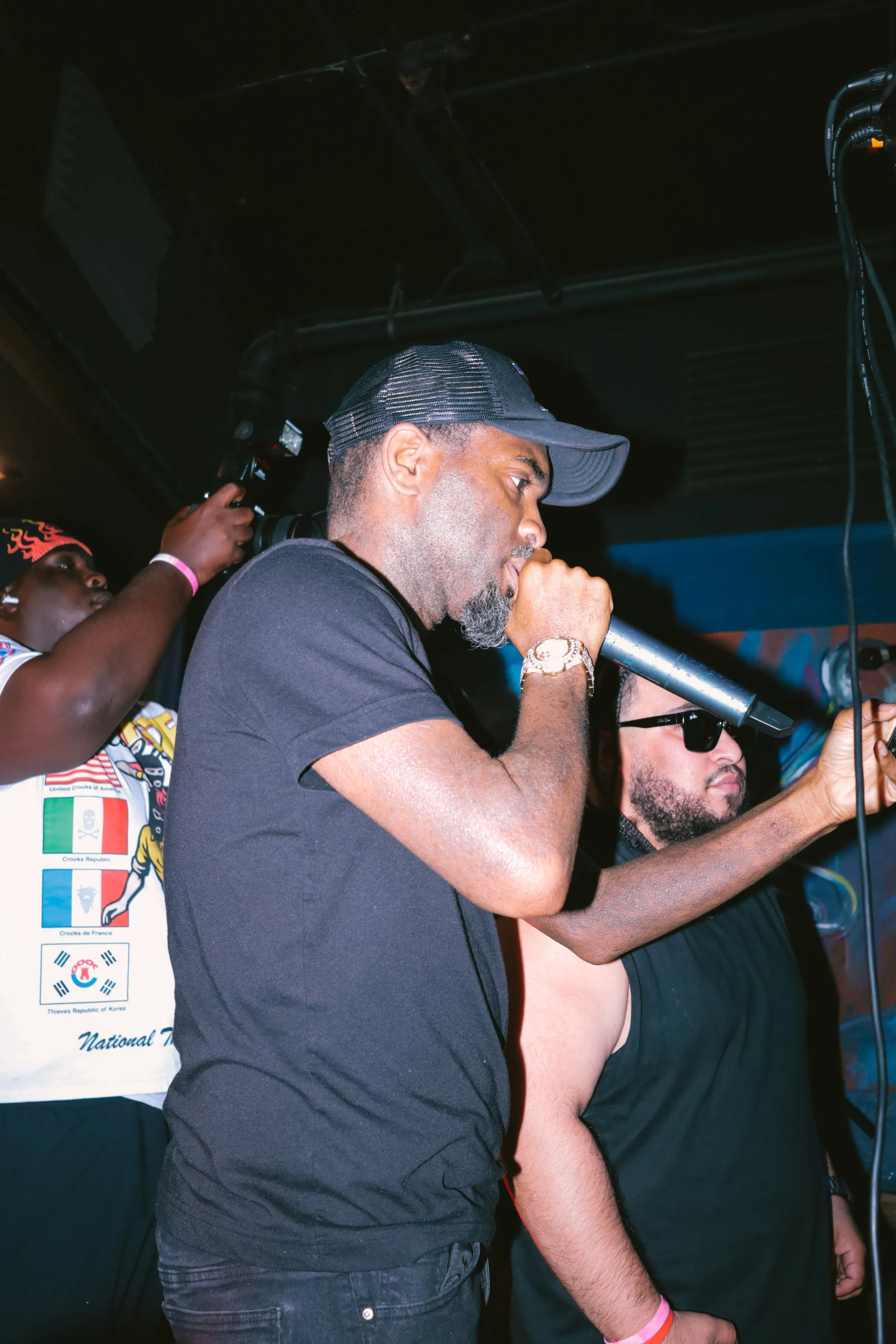 Three men are in a dark room, with one using a video camera, and another speaking into a microphone. The man with the microphone is wearing a gray cap, a black T-shirt, and a gold watch. The third man is wearing sunglasses and a black shirt.