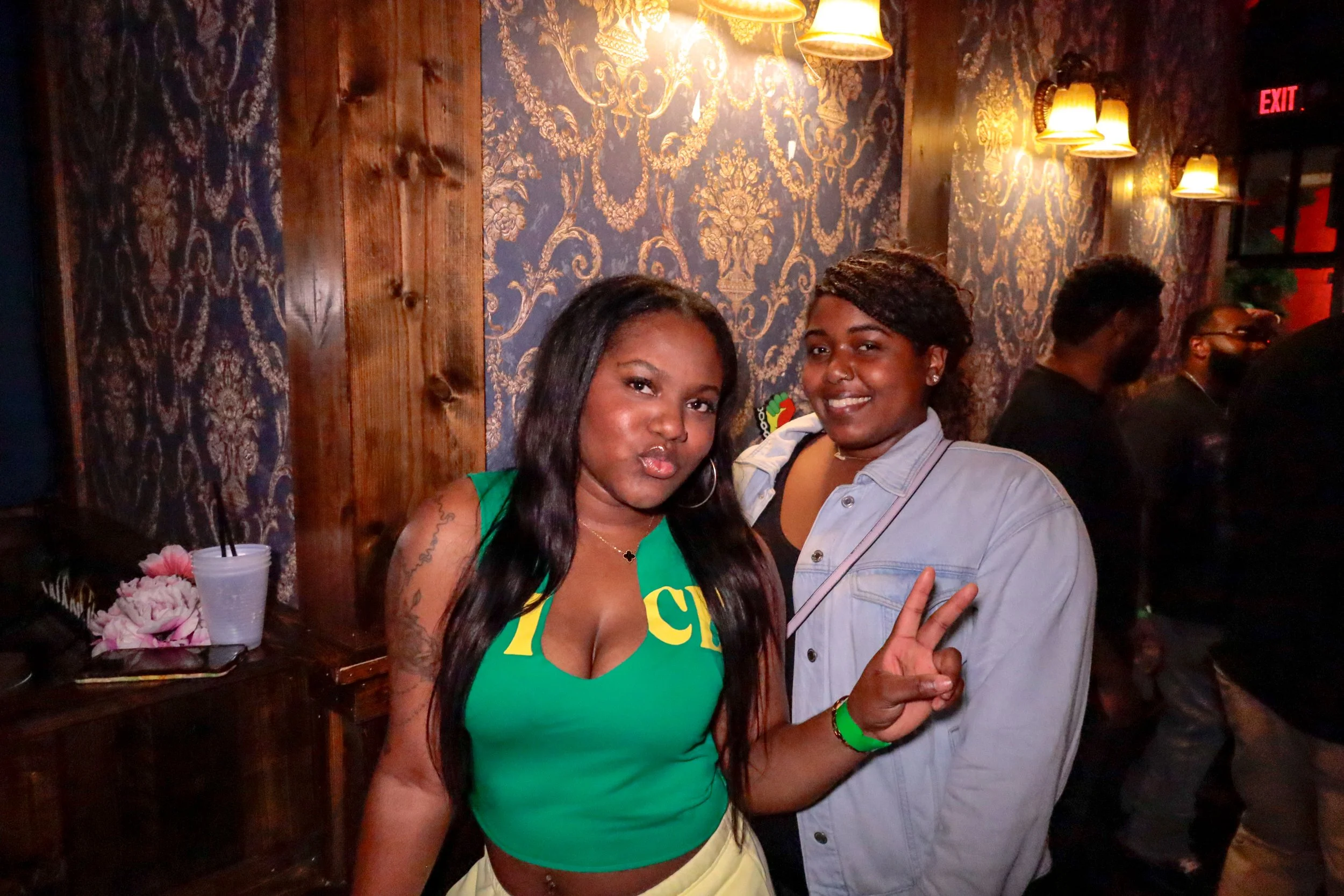 Two women posing for a photo in a dimly lit room with ornate wallpaper and wooden wall panels. The woman on the left wears a green shirt and makes a peace sign, while the woman on the right wears a light denim shirt. Other people are visible in the b