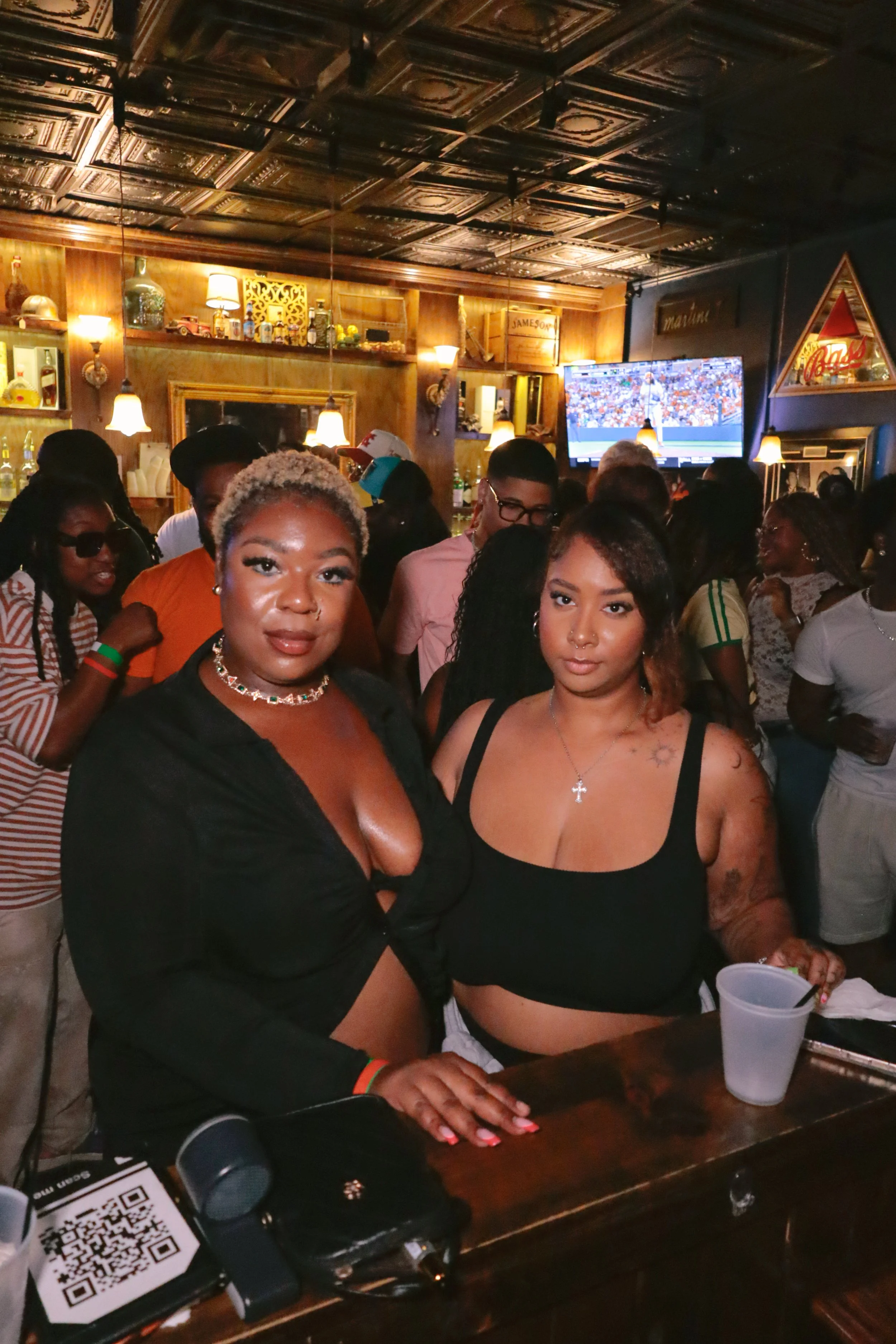 Two women stand at a bar or table in a crowded, warmly lit bar or pub, with many people in the background. One woman has short, platinum blonde hair and wears a black top with jewelry; the other has dark brown hair, tattoos, and wears a black tank to