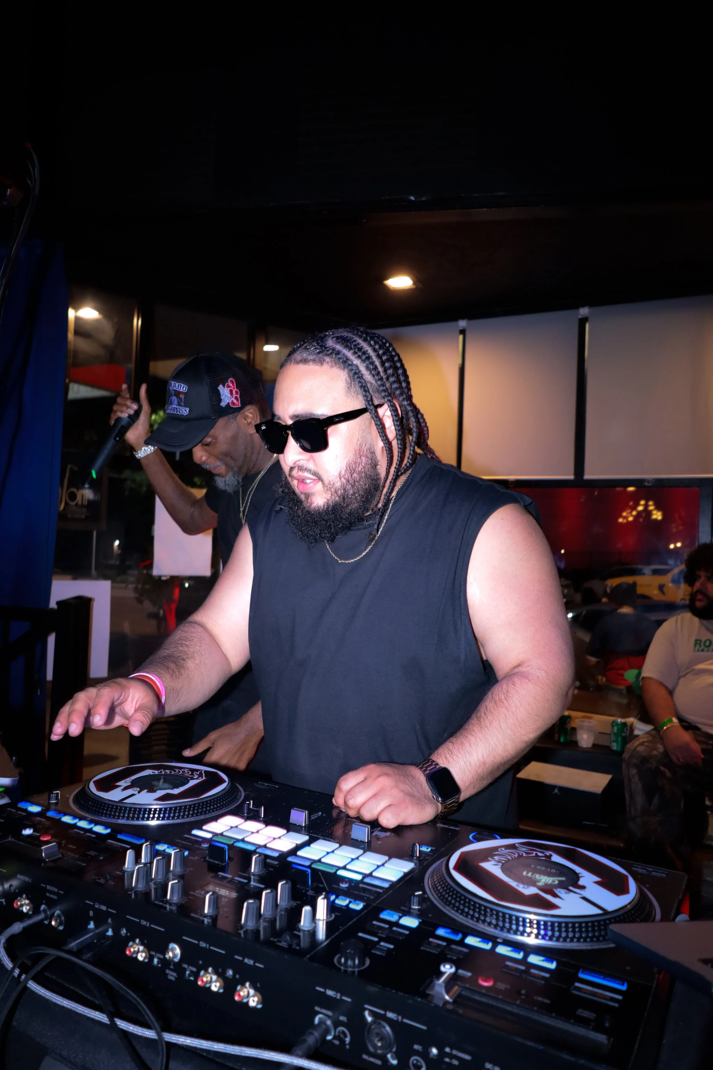 A DJ with braided hair, sunglasses, and a sleeveless shirt is playing music at a DJ booth in a club or party setting. There are people and colorful lights in the background.