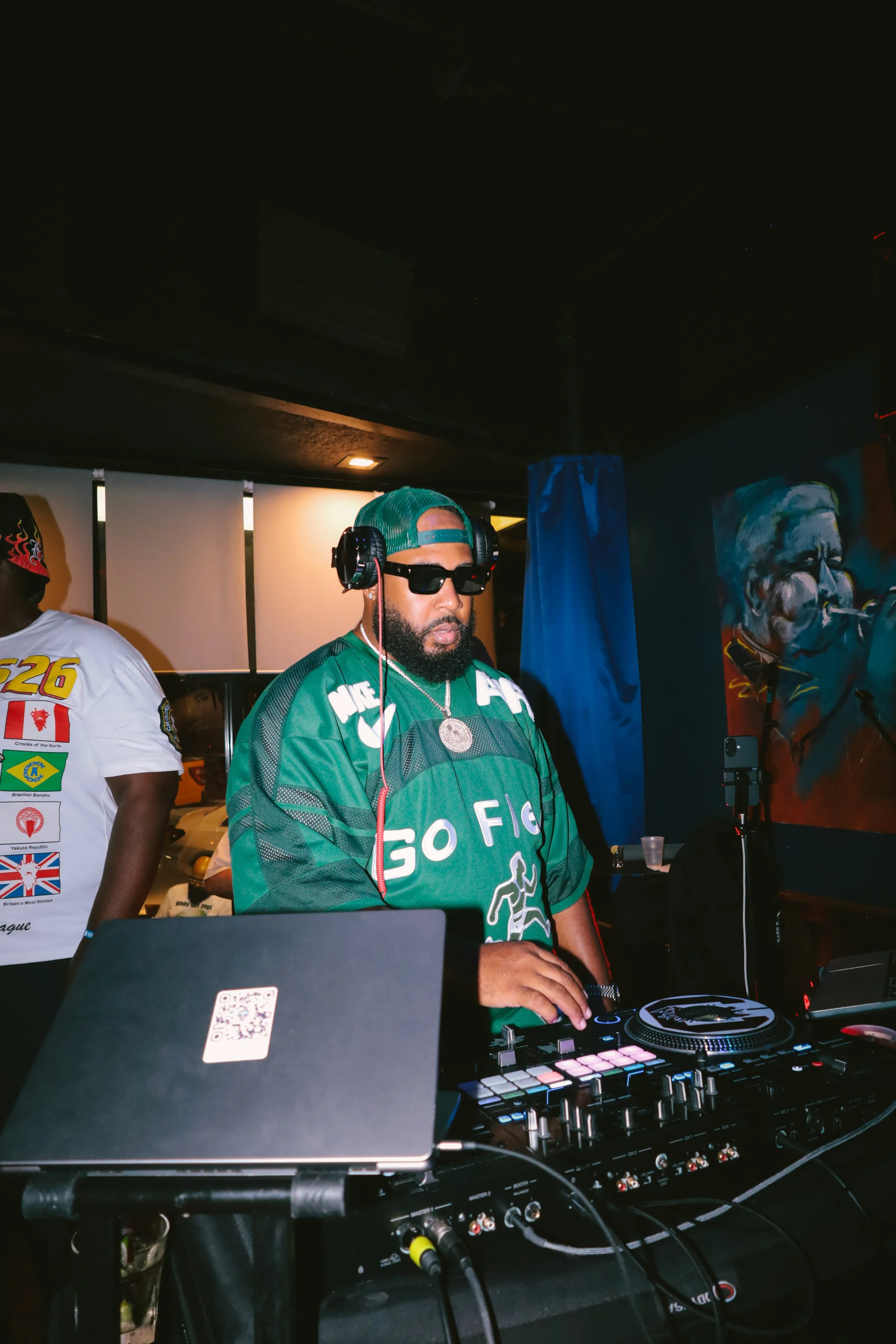 A DJ wearing sunglasses, a green cap, and a green jacket with white and black accents is playing music at a club or event. He is using DJ equipment, including a turntable and mixer, with various wires connected. Another person wearing a white T-shirt