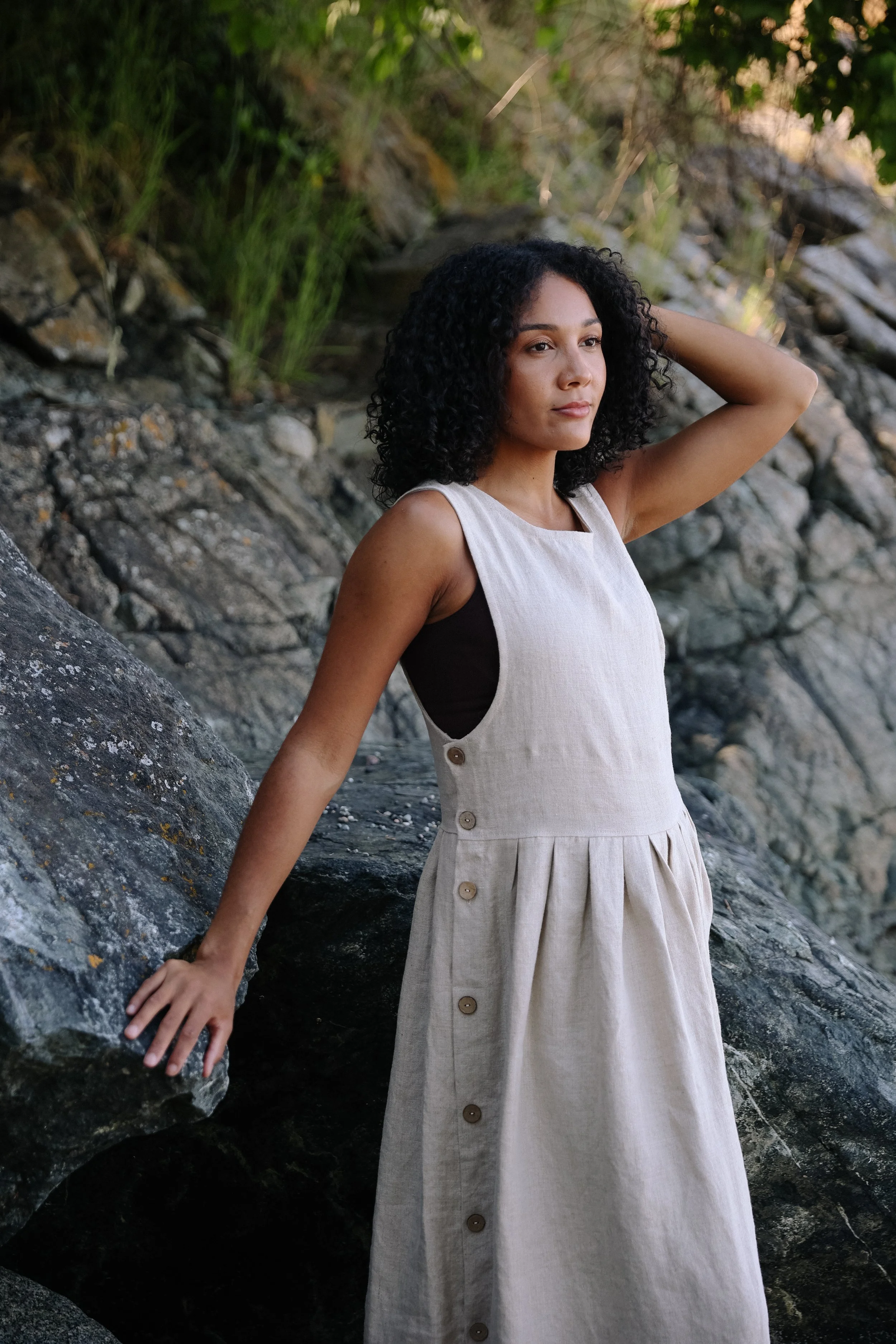 A woman with dark curly hair is standing outdoors against a rocky and grassy background. She is wearing a sleeveless light-colored dress with wooden buttons, and her left arm is raised with her hand resting on her head while her right arm is extended