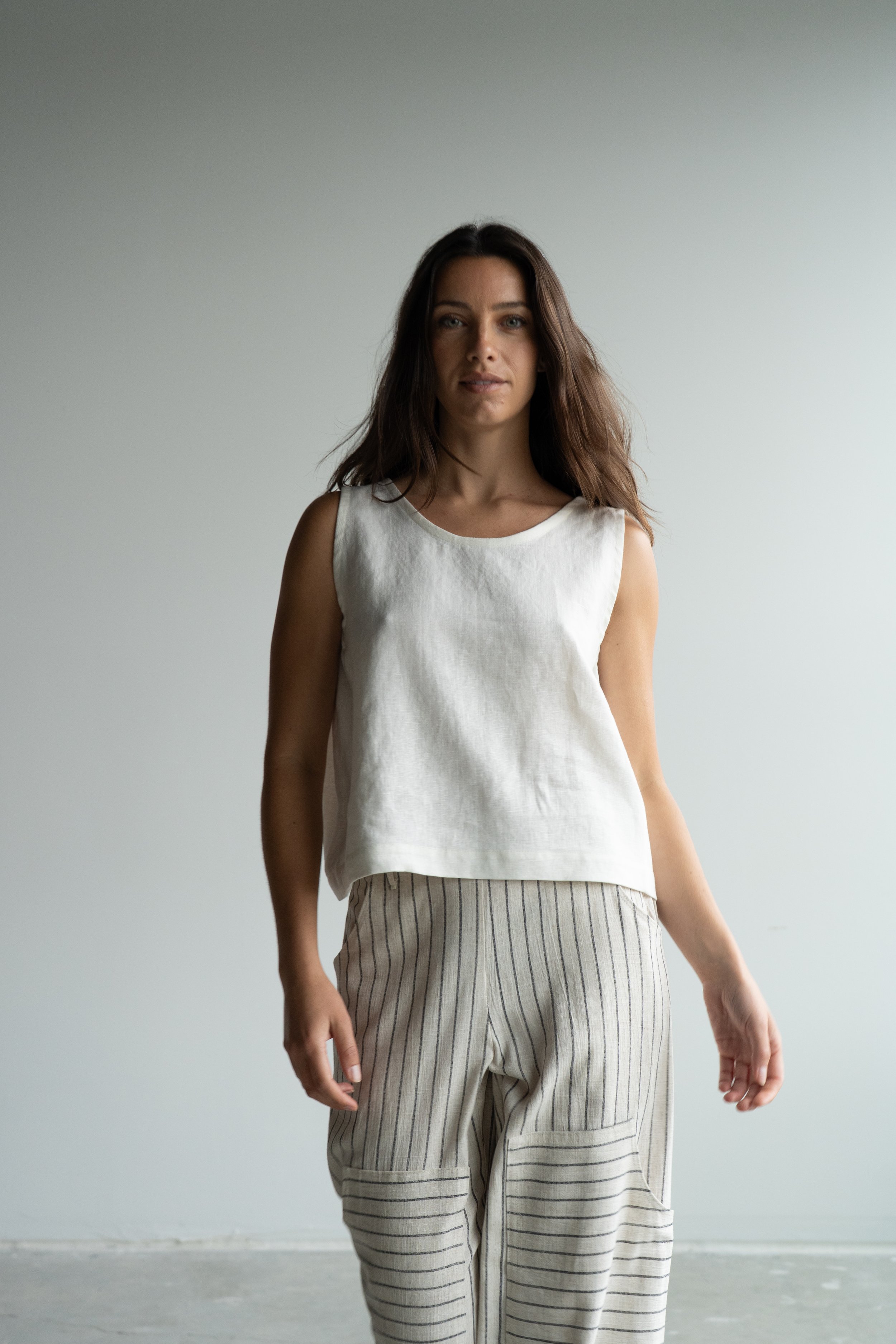 A woman with long brown hair standing in front of a plain white wall, wearing a sleeveless white top and striped linen pants.