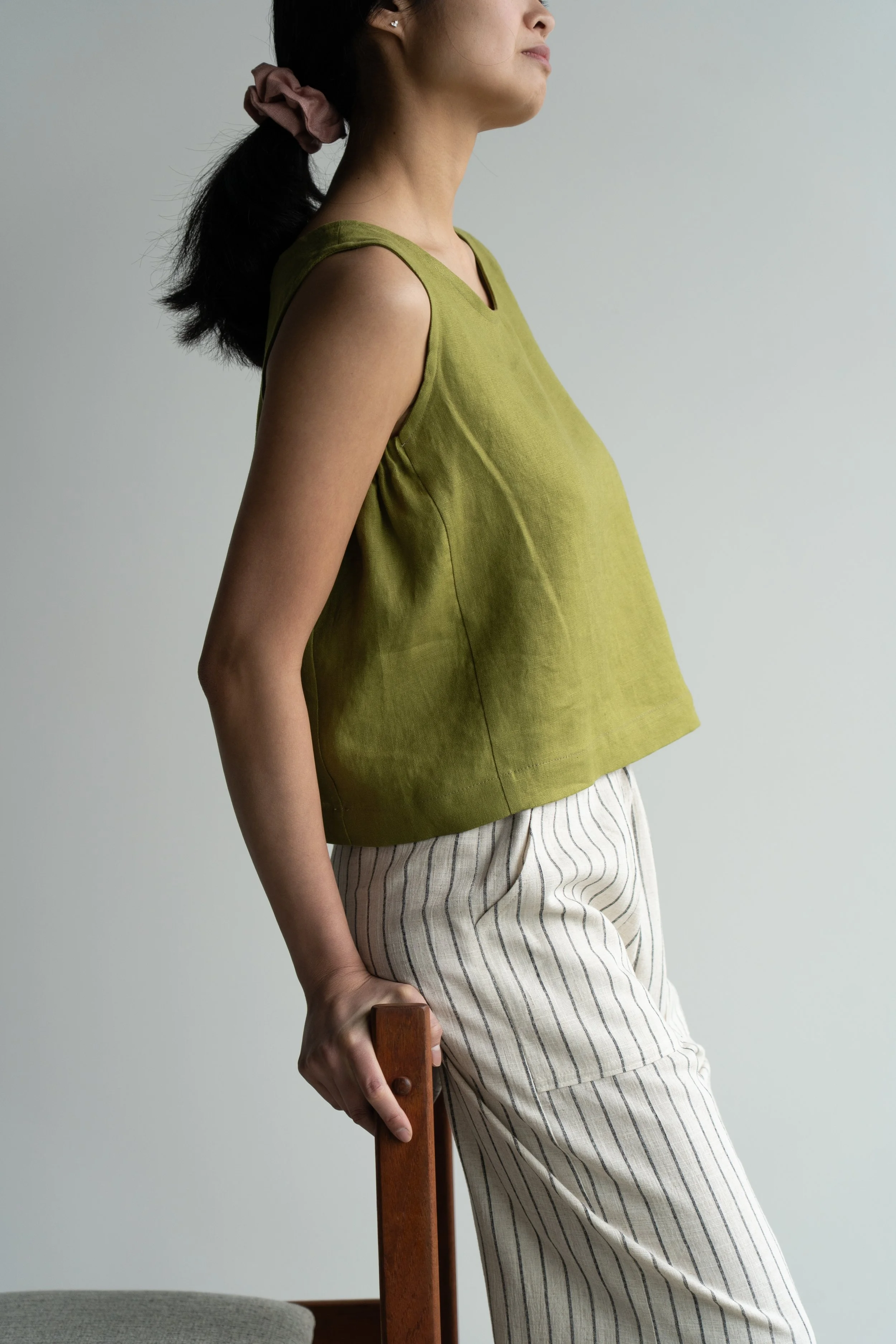 A woman standing sideways, wearing a green linen sleeveless top and striped pants, holding the back of a chair with her hand.