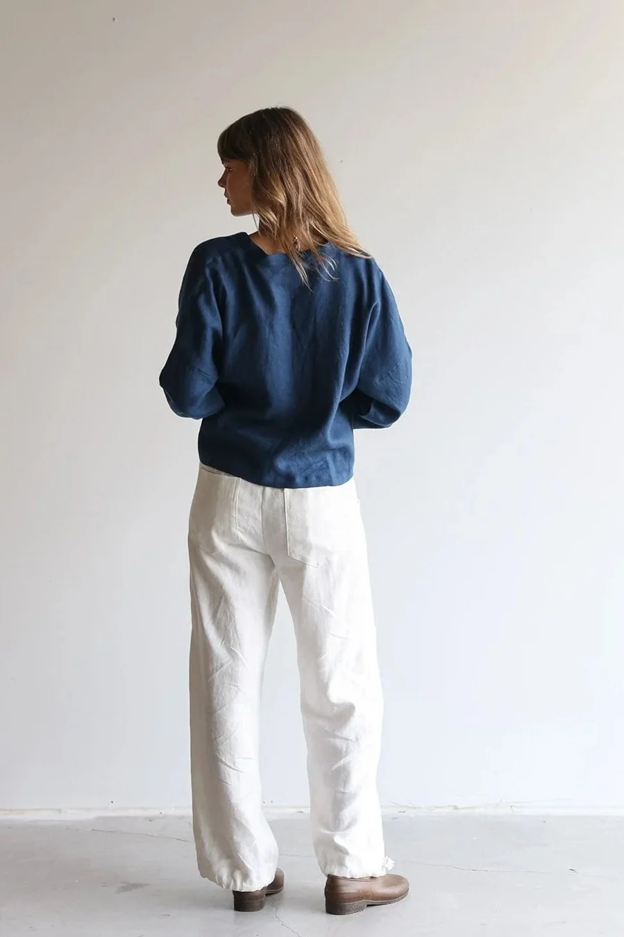 Back view of a woman wearing a linen wrap top in blue looking left with white linen pants