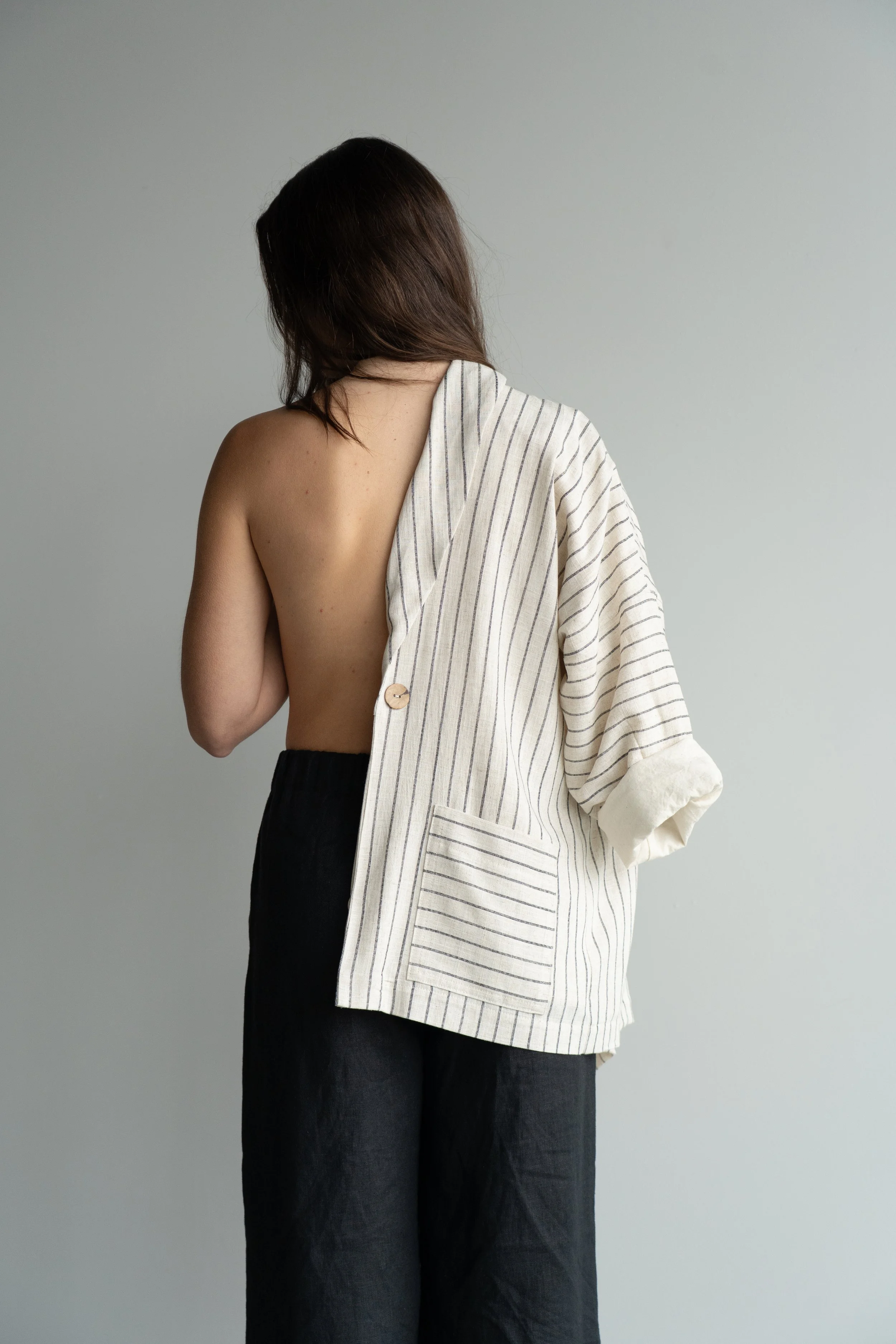 Person with shoulder-length dark hair, partially turned away, wearing black pants and a cream-colored striped blazer, with one side off the shoulder showing bare skin, standing against a plain light gray background.