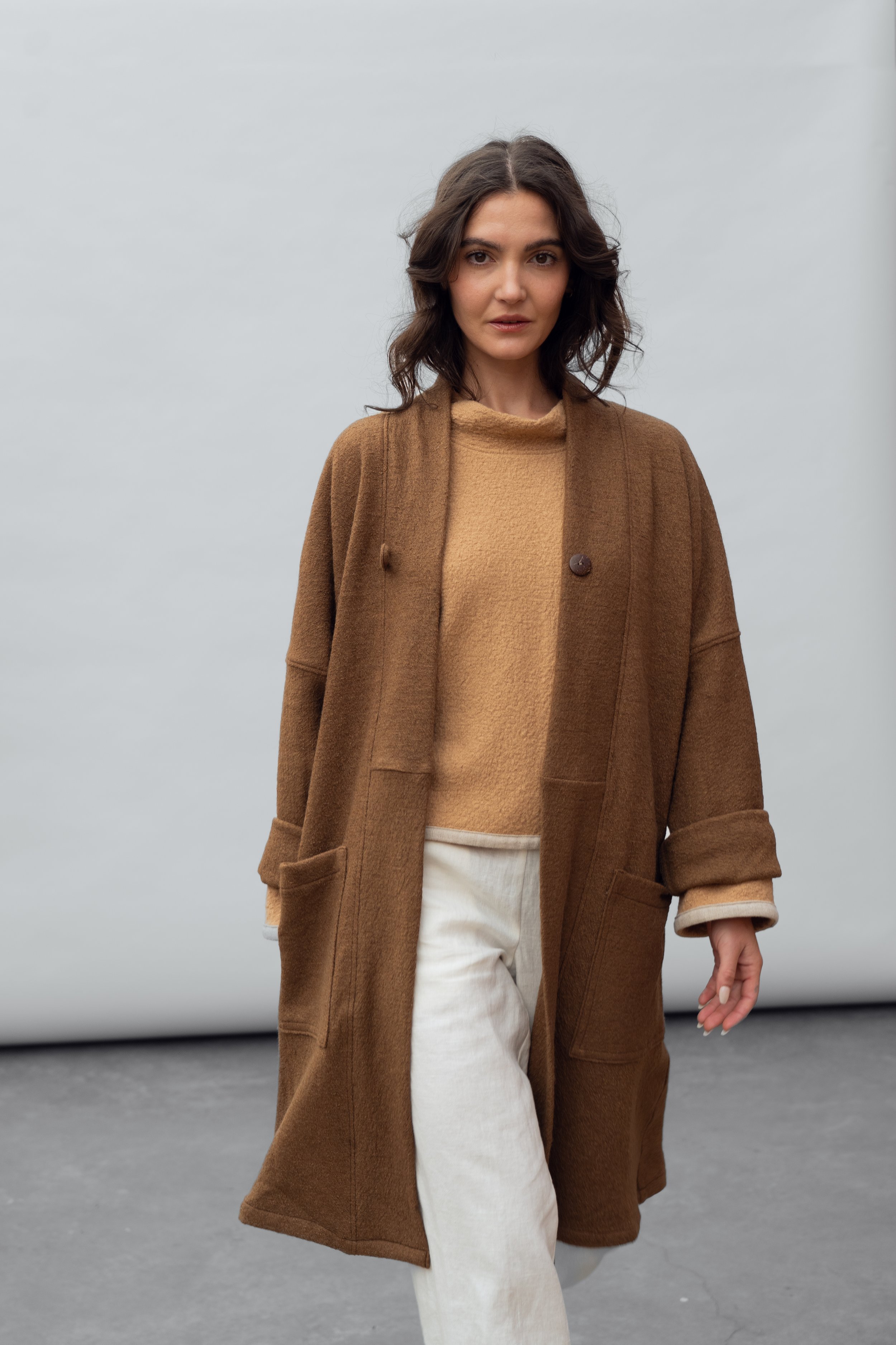 Woman with dark hair wearing a tan sweater, oversized brown coat, and light-colored pants standing against a plain background.