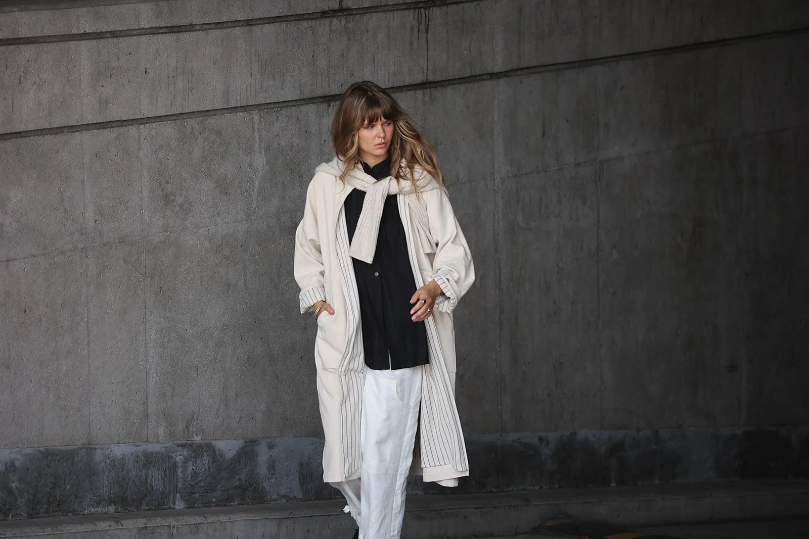 Overhead view of a woman wearing a canvas linen trench coat in Sand walking with hand in pocket in front of a concrete wall