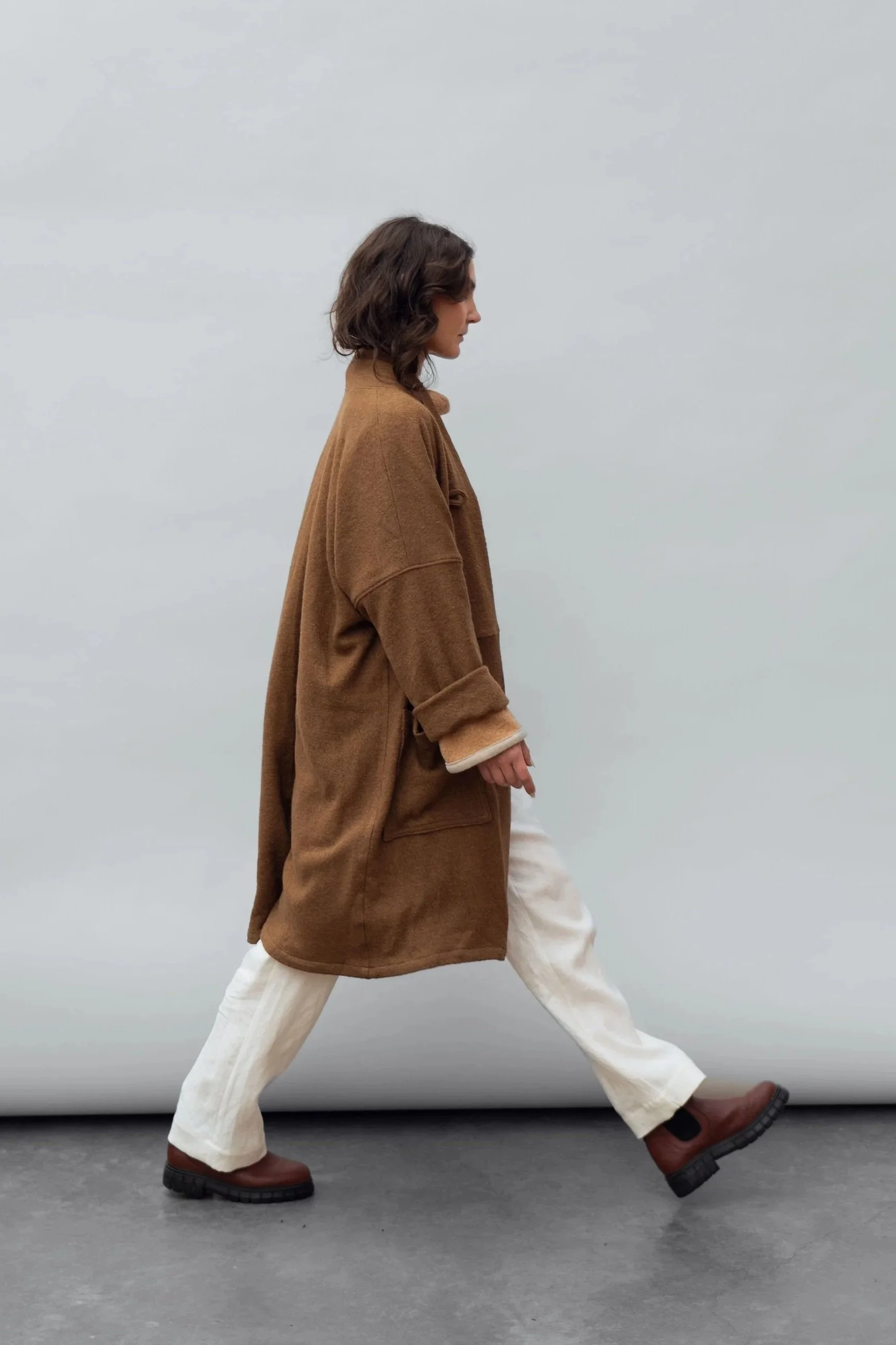 Side view of a woman wearing a wool coat in Bark and white pants walking to the right